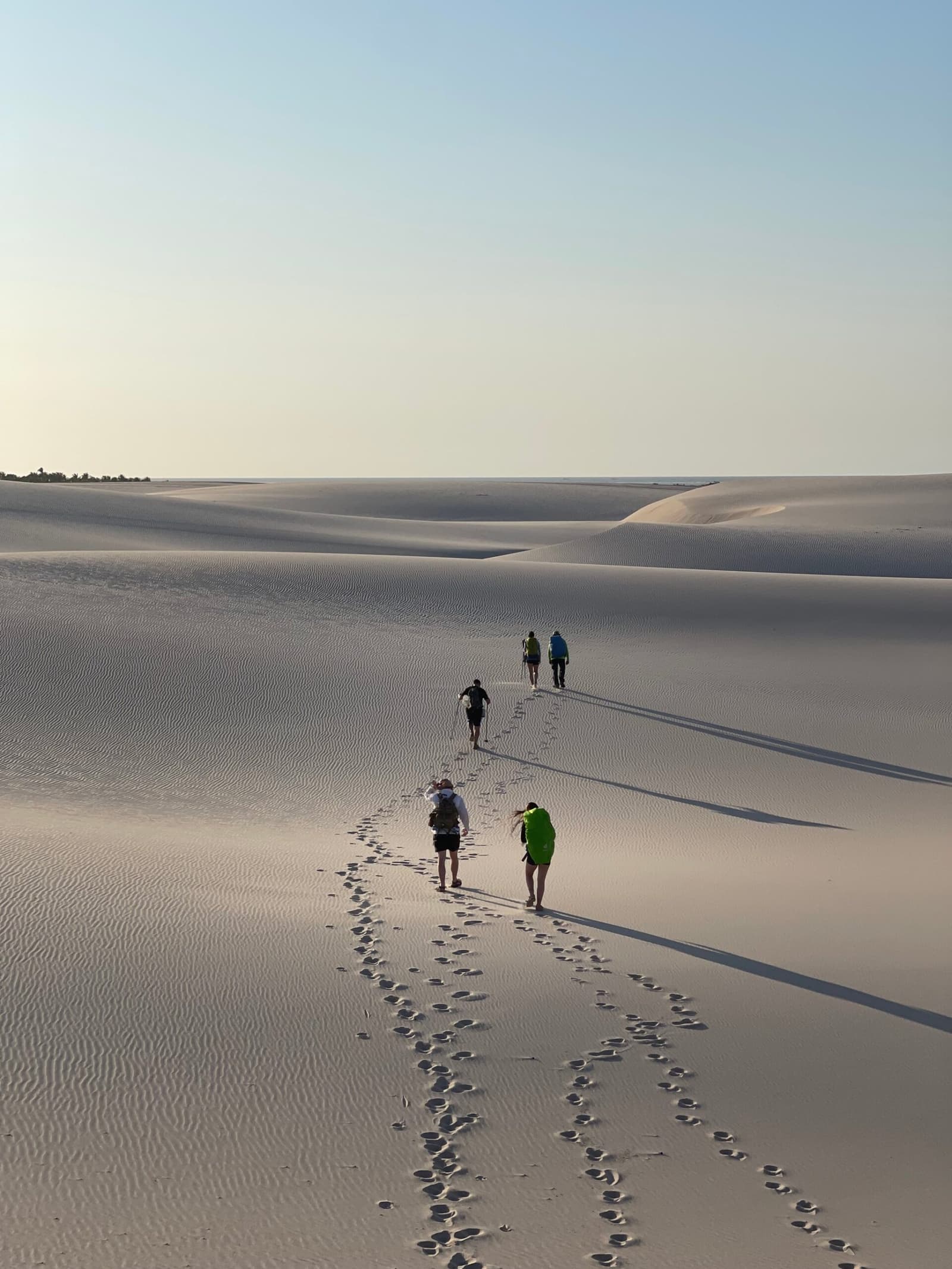 Four people, one set of footprints, and a whole lot of sand to cover. Four hikers with backpacks walking across vast white sand dunes on a Lençóis Maranhenses hike, their footprints tracing a long trail behind them under a pale morning sky