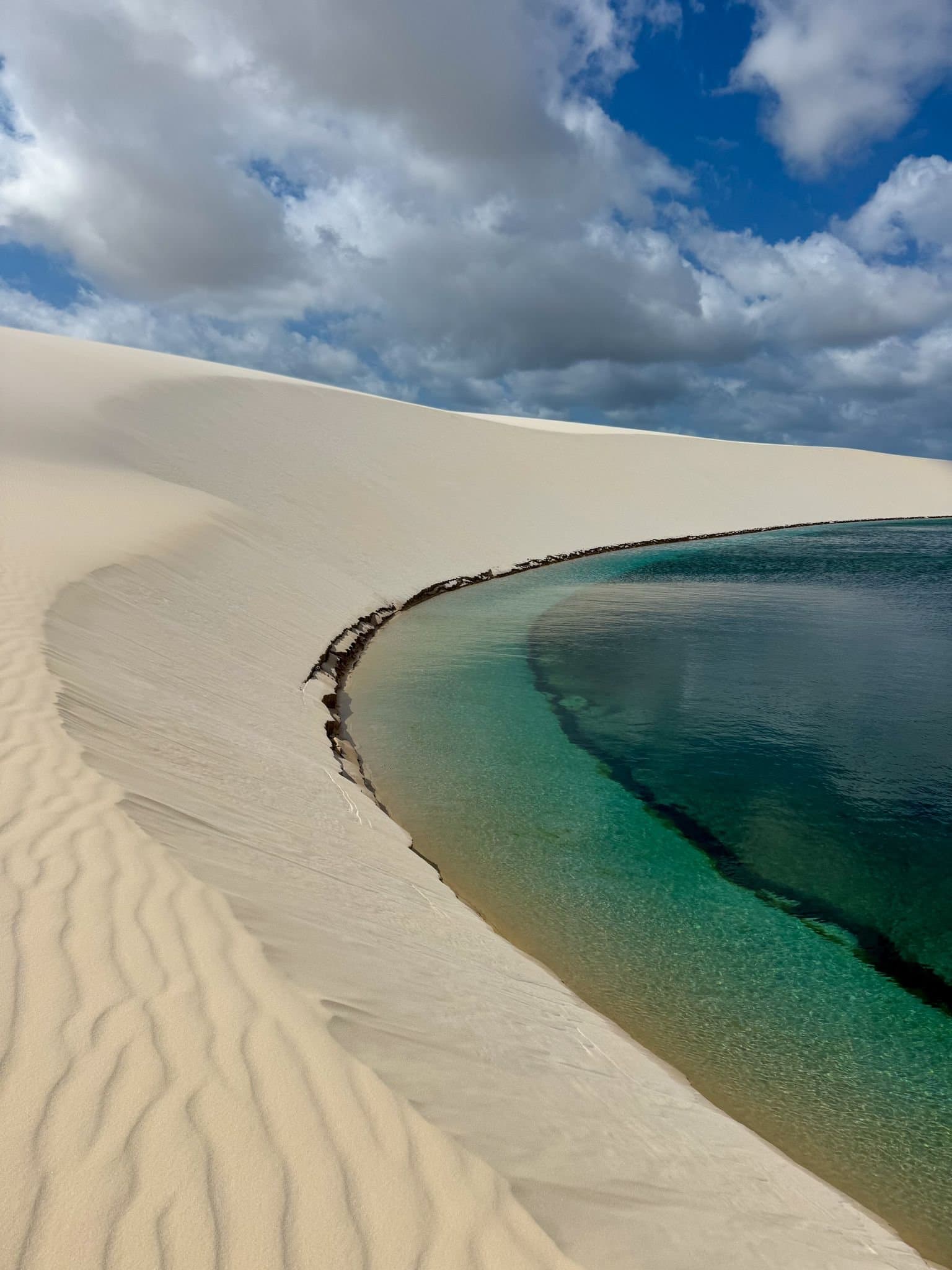 Sand this white, water this clear, Lençóis Maranhenses keeps its promises. A crescent-shaped turquoise lagoon nestled against a sweeping white sand dune under a partly cloudy blue sky in Lençóis Maranhenses Santo Amaro, with the shallow shoreline fading from clear aqua to deep teal
