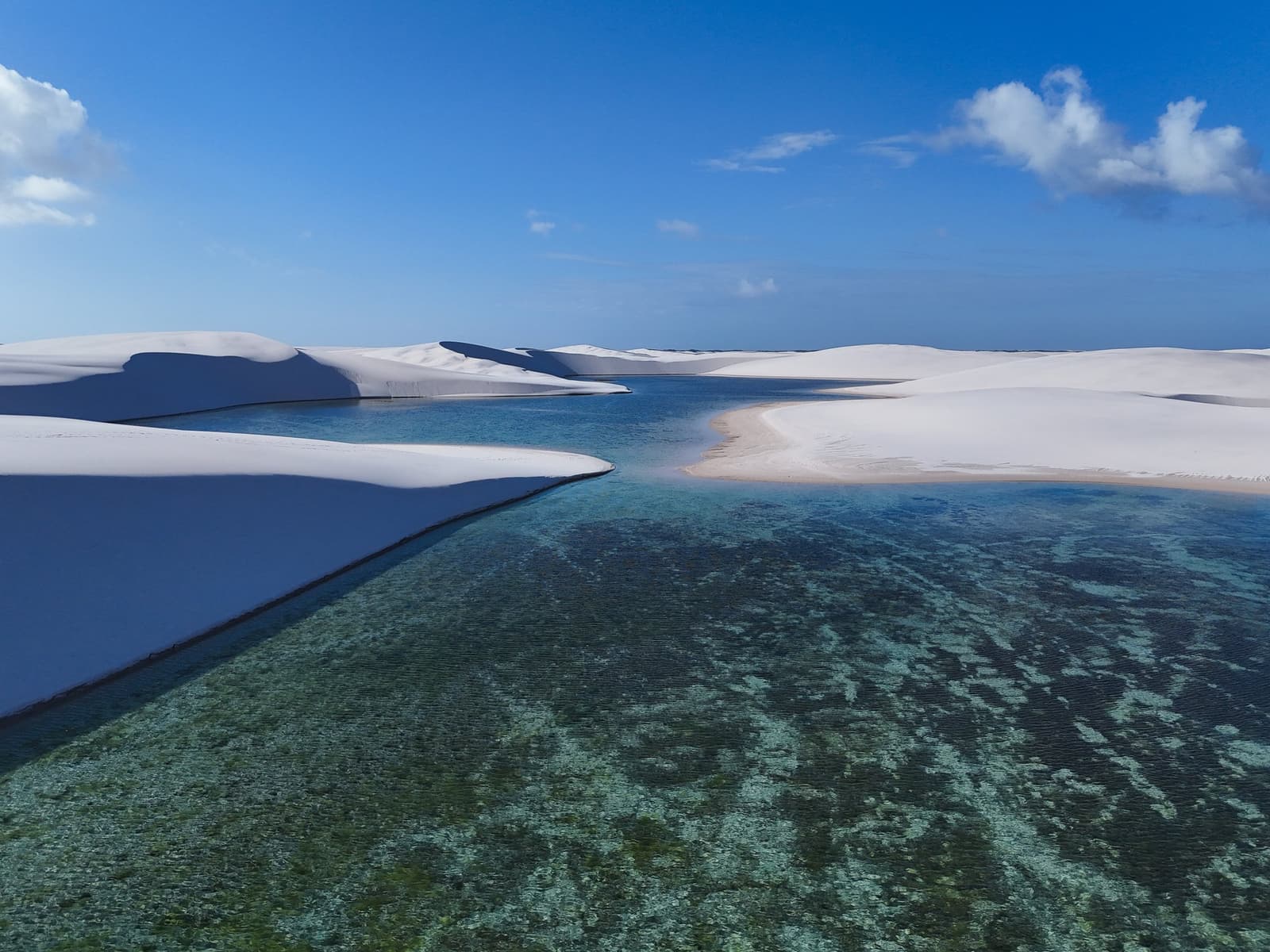 Half desert, half lake, and somehow both at once. Aerial view of a crystal-clear green freshwater lagoon nestled between sweeping white sand dunes under a blue sky in the Lençóis Maranhenses trek from Atins route