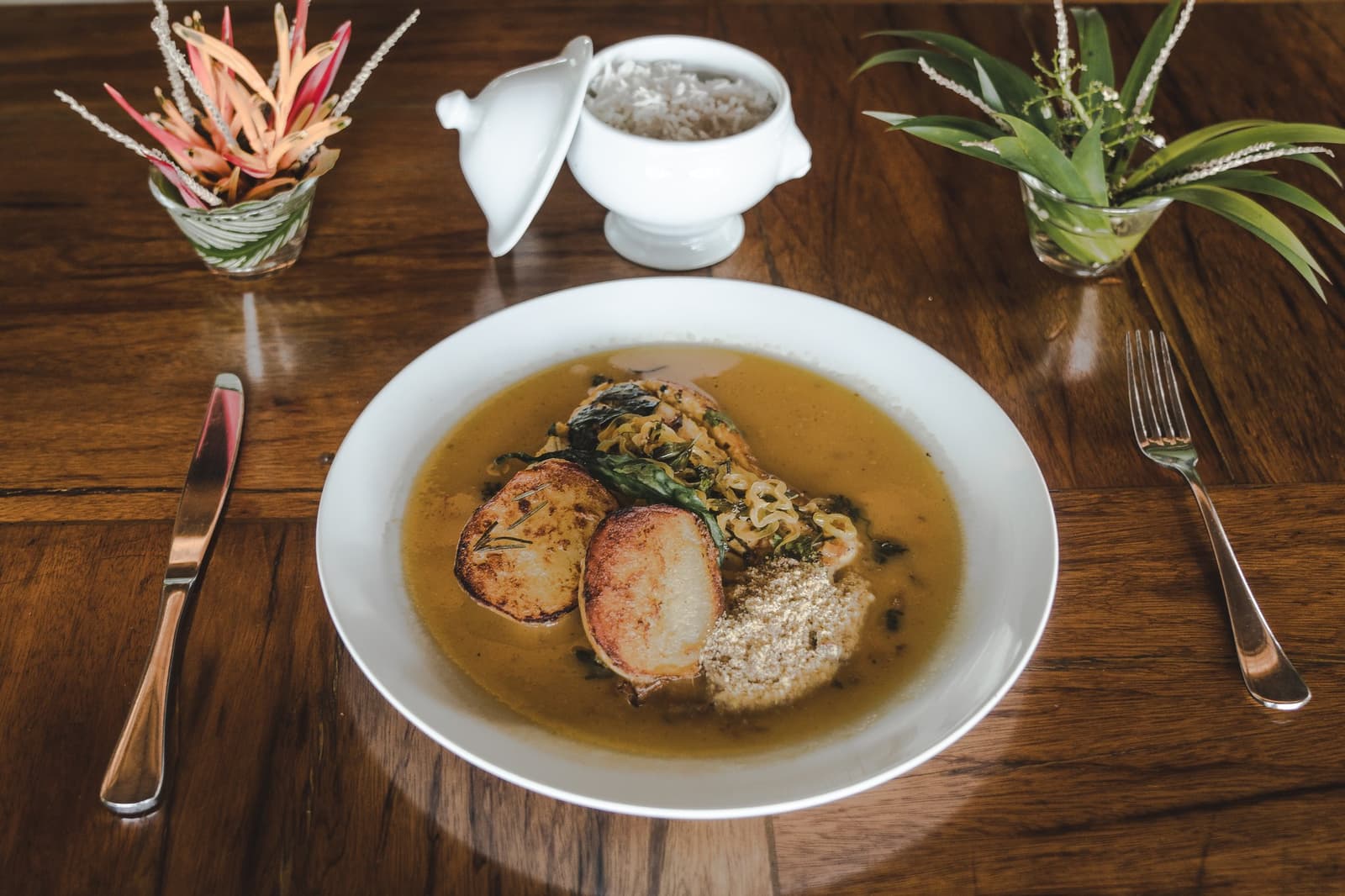 Lodge cooking that takes local ingredients seriously, and it shows. A white bowl of braised meat in golden broth with sautéed greens, toasted bread rounds, and cassava flour, served at the Mirante do Gavião lodge restaurant alongside a white tureen of rice and tropical plant arrangements on a polished timber table
