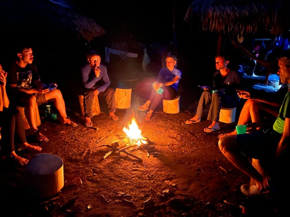 Good conversation travels further by firelight. Everyone leans in. A group of travelers on a Pico da Neblina Expedition seated on log stumps in a circle around a small campfire at night, beneath a thatched shelter in the Amazon rainforest