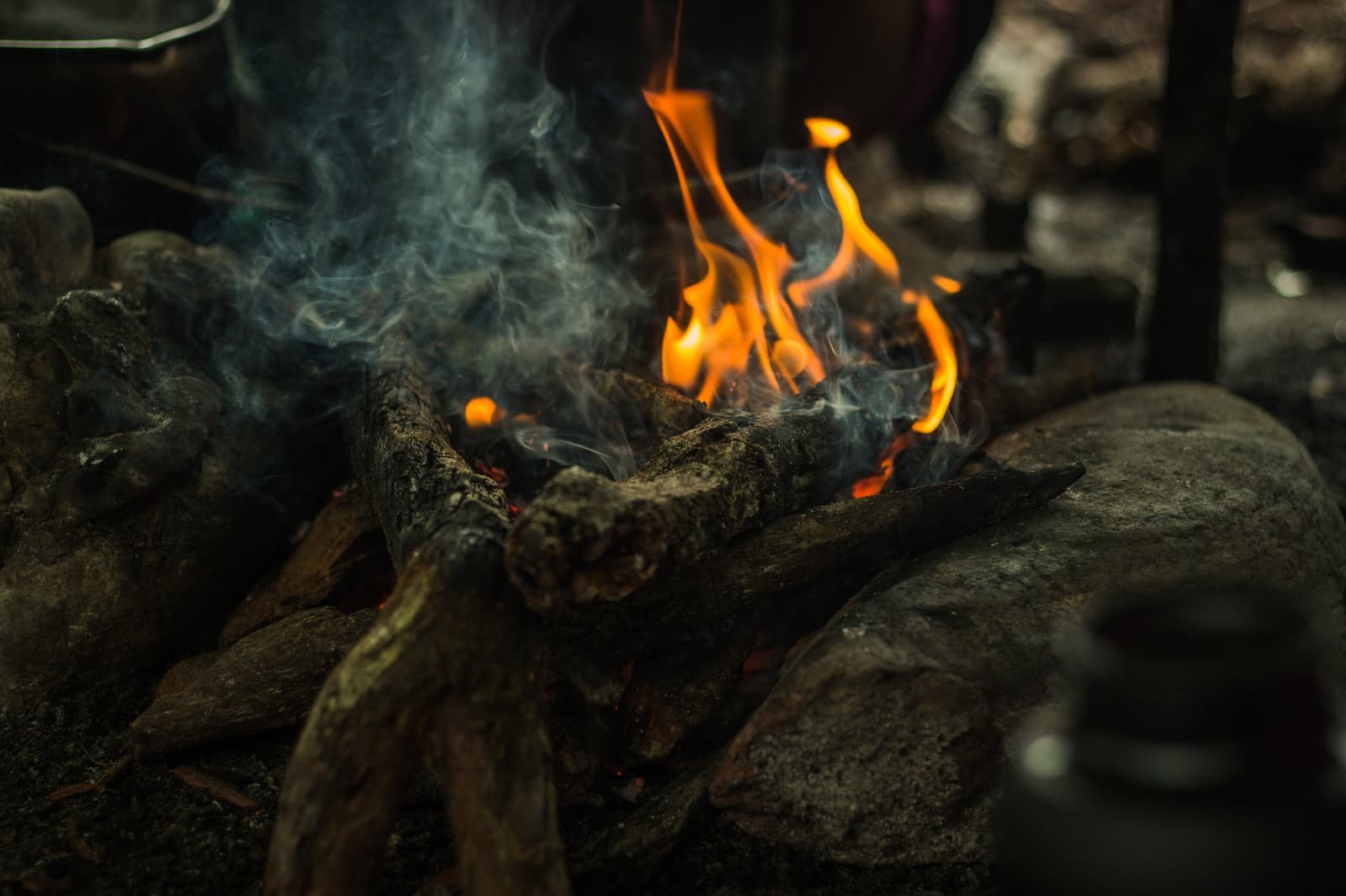 Fire started, stones set, dinner earning its keep in the dark. Close-up of a wood fire burning between river stones on a Pico da Neblina Expedition camp, orange flames rising through smoke above charred logs