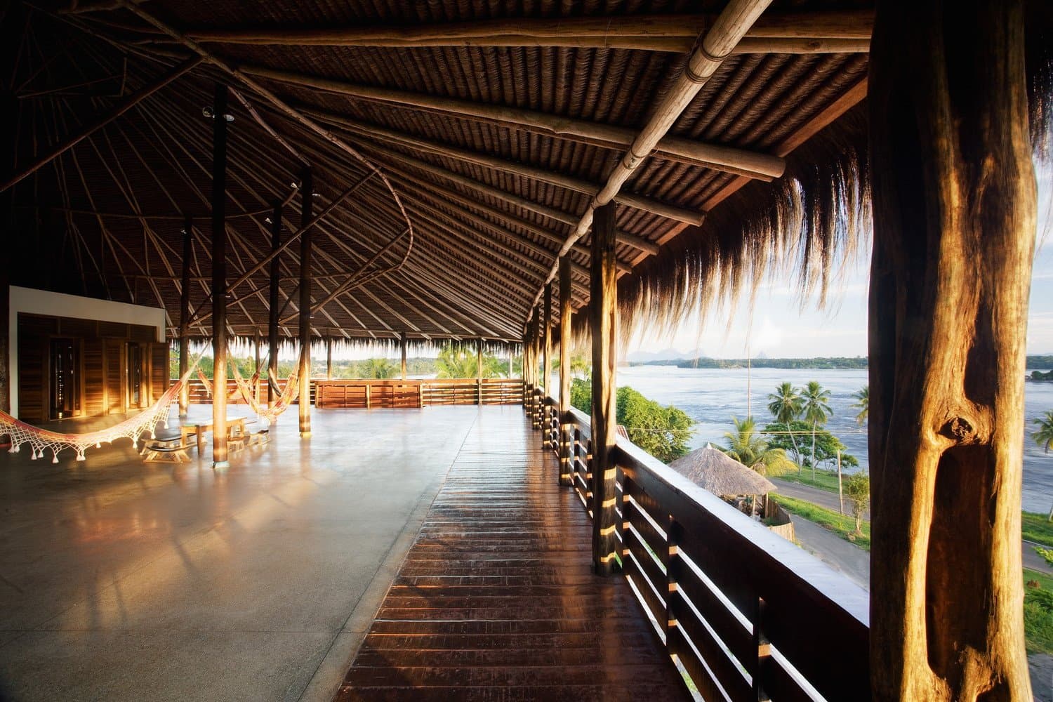 Hammocks, warm light, and a river that makes the packing feel very far away. Photo: Daniel Ducci Open thatched-roof terrace of a riverside lodge on the Pico da Neblina Expedition, with hammocks strung between wooden posts and a wide view over a broad river lined with palm trees at golden hour