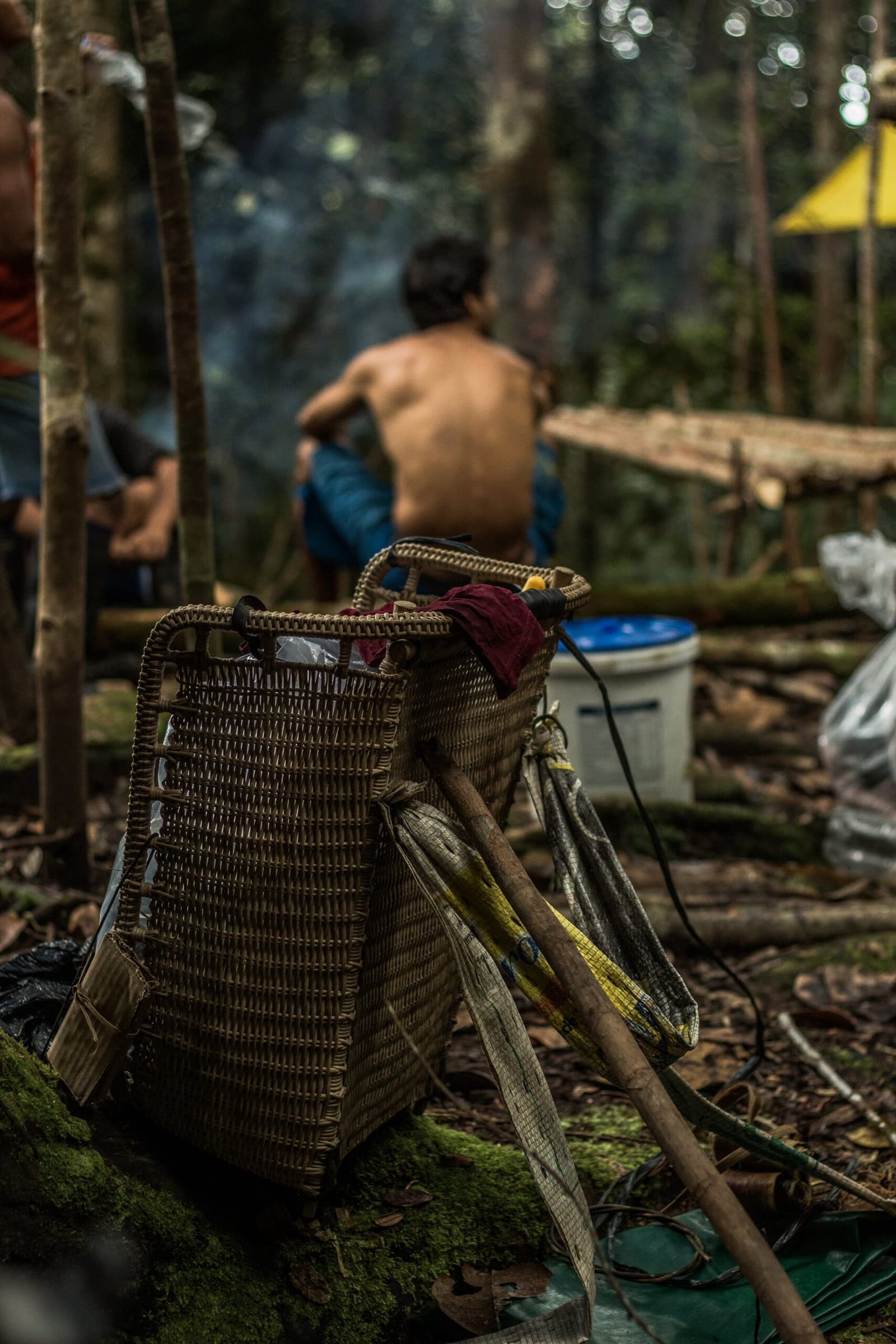 Packed, unpacked, repacked, the basket earns its keep every single day. A traditional woven wicker backpack basket rests against a mossy root on the forest floor during a Pico da Neblina Expedition camp stop, with a shirtless man sitting by a fire in the blurred background among tall rainforest trees