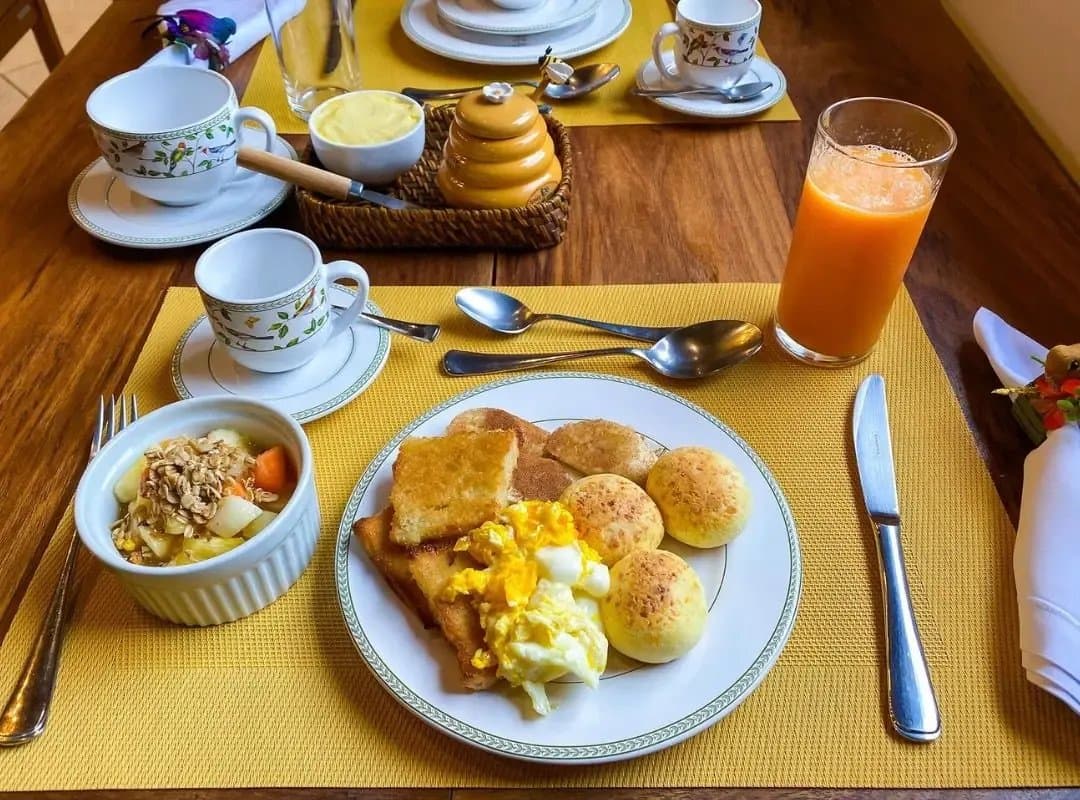 Pão de queijo at breakfast. Somehow, the bar is set before 8 AM. A southern pantanal safari lodge breakfast plate with scrambled eggs, toast, and pão de queijo alongside fresh orange juice, granola with fruit, honey, and floral-patterned coffee cups on a yellow placemat