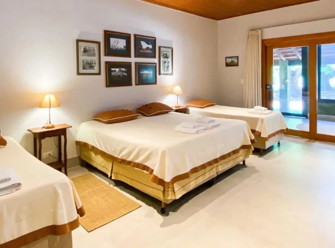 Clean sheets, warm light, and greenery just outside the door. Sleep well, the anteaters start early. Triple room at a southern pantanal safari lodge, with three beds dressed in cream and caramel linens, warm bedside lamps, framed black-and-white photographs on the wall, and sliding glass doors opening to garden greenery