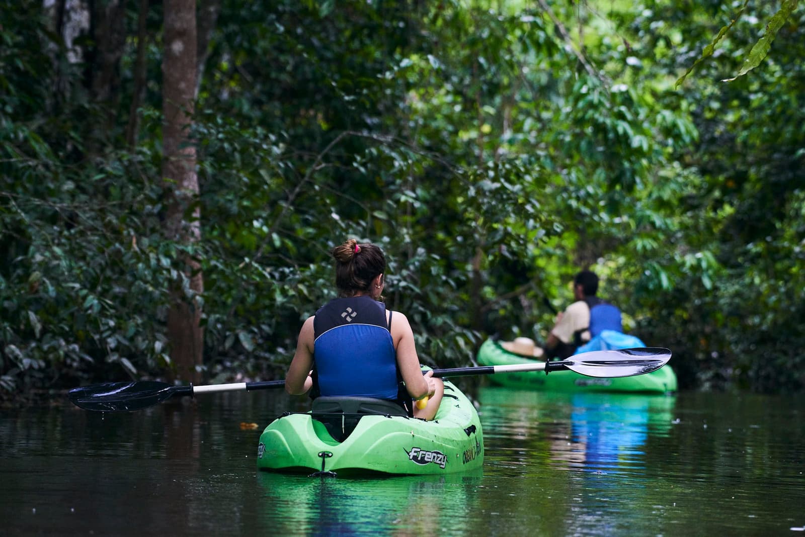 Quiet water, thick canopy, no agenda. Just you and the forest deciding the pace. Photo: JUKAPAZ Two kayakers paddling green sit-on-top kayaks through a narrow, dark-watered channel lined with dense rainforest canopy on a tucano amazon river cruise igapó excursion