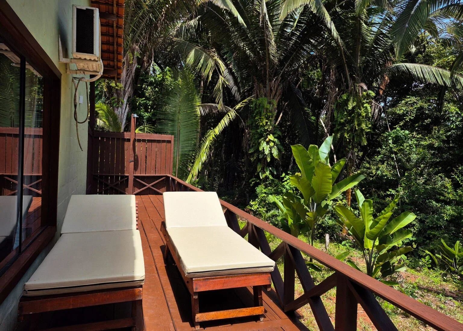 Two chairs, zero agenda, and the forest doing its thing right outside. Two cushioned wooden sun loungers on a private timber deck at an Amazon boutique lodge, surrounded by dense tropical vegetation including palm trees and banana plants
