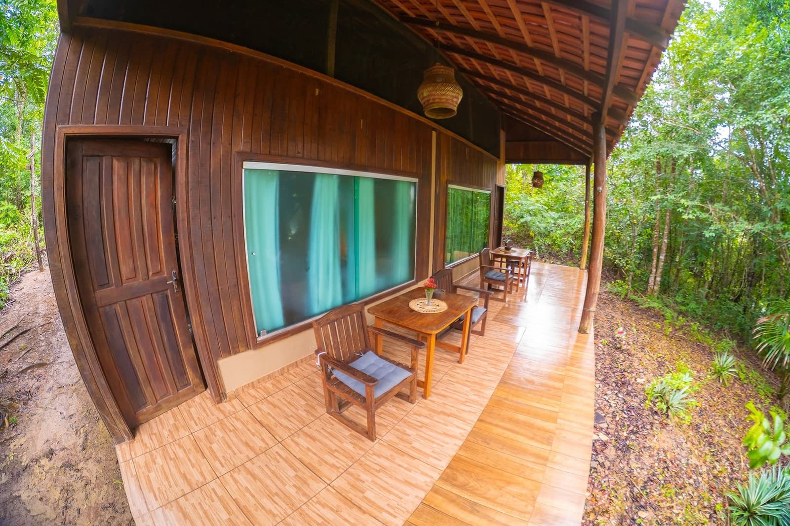 Your own front-row seat to the forest, no binoculars required. A covered timber veranda at Amazon Turtle Lodge with wooden chairs, cushioned seats, and small dining tables stretching along a dark-wood bungalow, surrounded by dense rainforest vegetation
