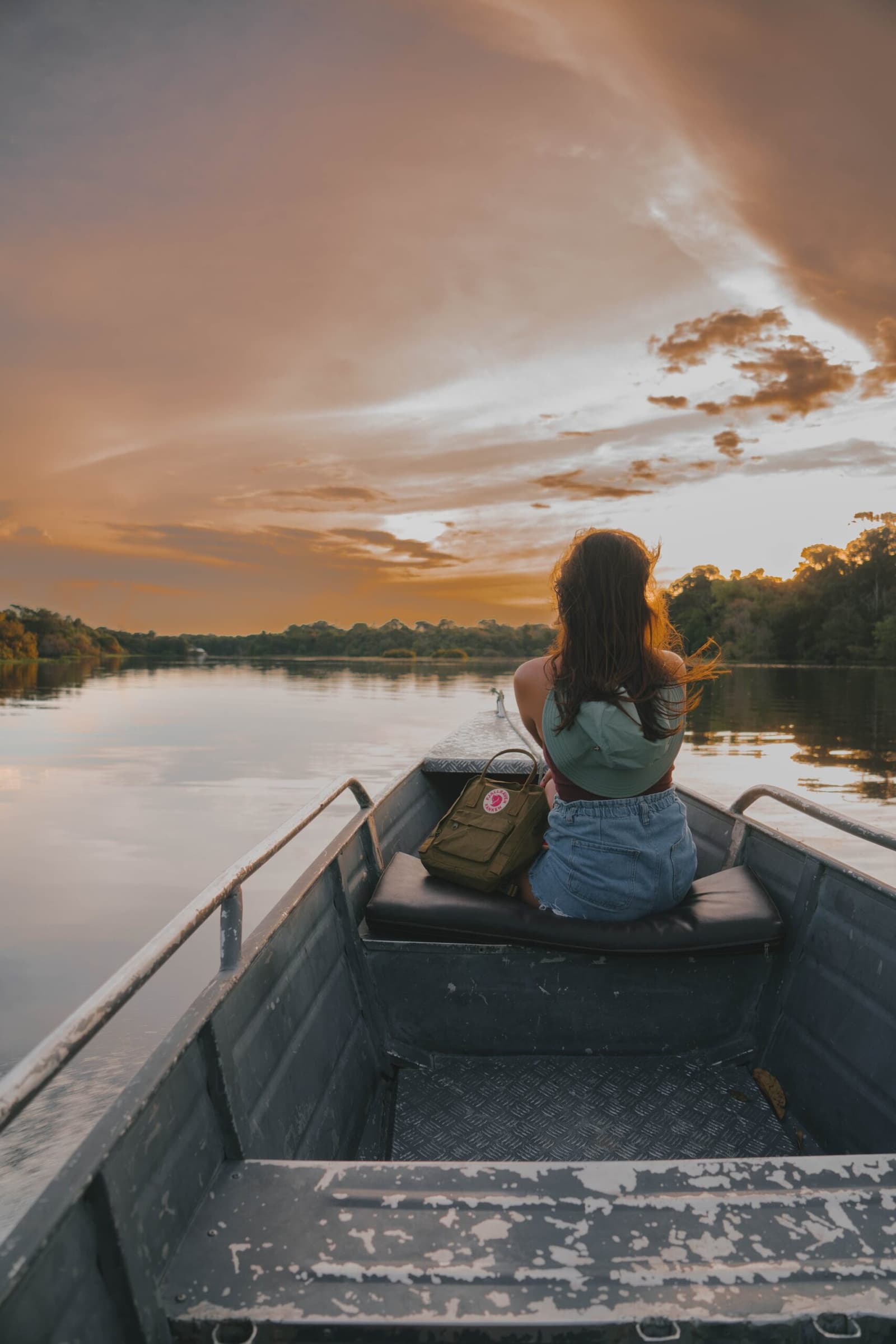 Some sunsets just stop you mid-thought. The river can wait. A traveller sits at the bow of a metal river boat watching a vivid orange sunset over a calm, jungle-fringed waterway on an amazon turtle lodge rainforest trip