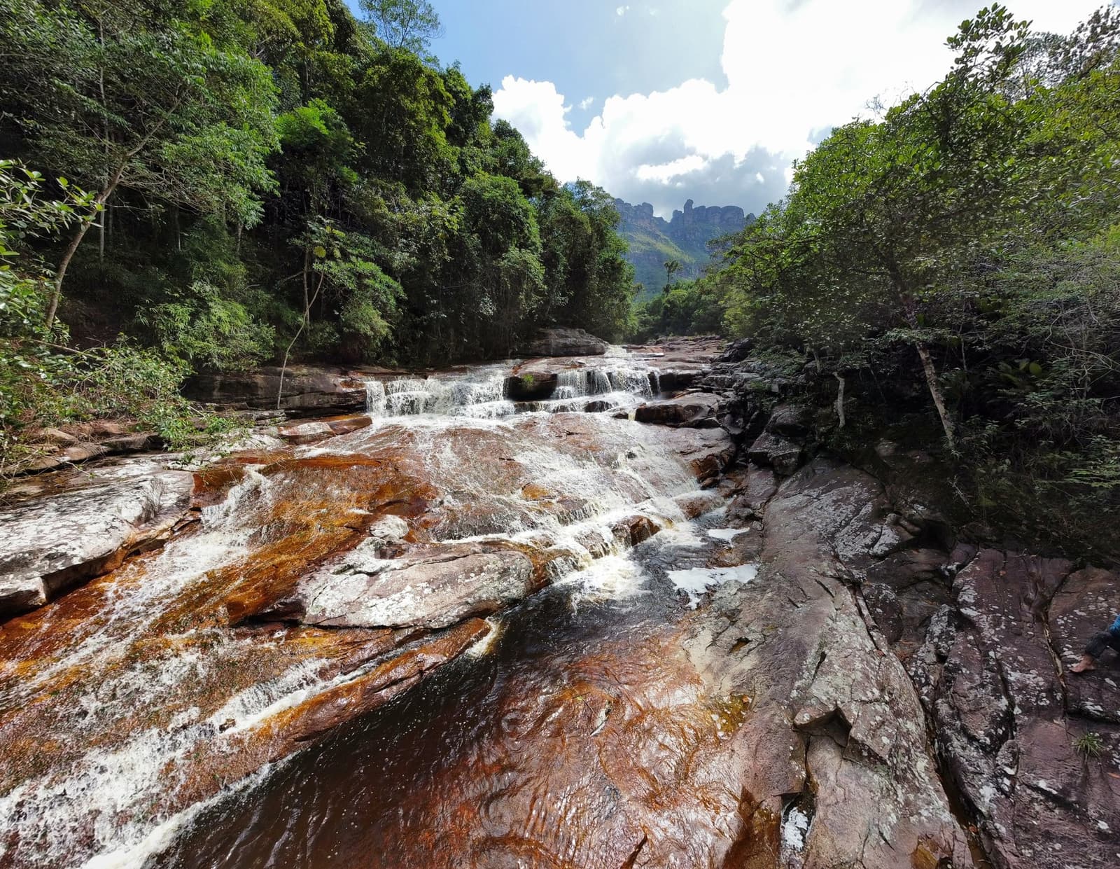 Iron-stained rock, cold water, bare feet. The Pati River has its own agenda. Clear water rushing over rust-tinted sandstone ledges in a tiered cascade on the Chapada Diamantina trek, with dense rainforest and jagged rock formations rising in the background