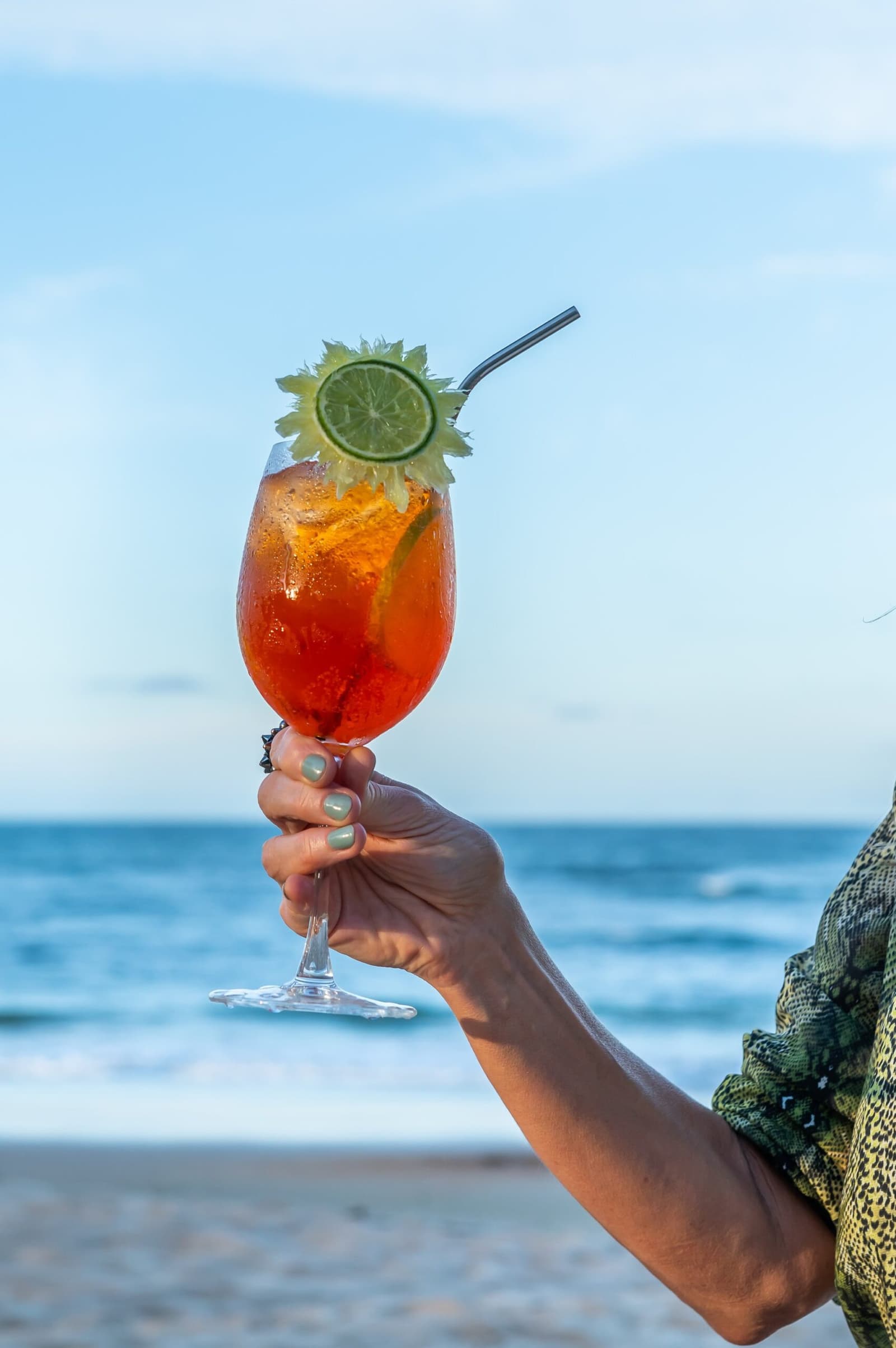 Earned it. A cold drink and the sea, nothing else required. A hand holds a large iced red cocktail garnished with a lime wheel and herbs, with the ocean horizon visible in the background on the Bahia coast