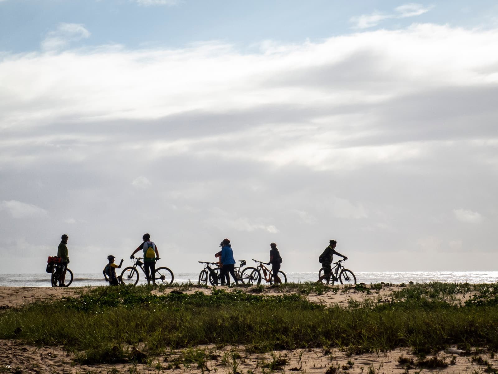 Six riders, one horizon, and the whole route still ahead. Group of cyclists with mountain bikes pausing on a sandy coastal dune covered in low vegetation, silhouetted against a bright cloudy sky on the cycling Bahia Cocoa Route