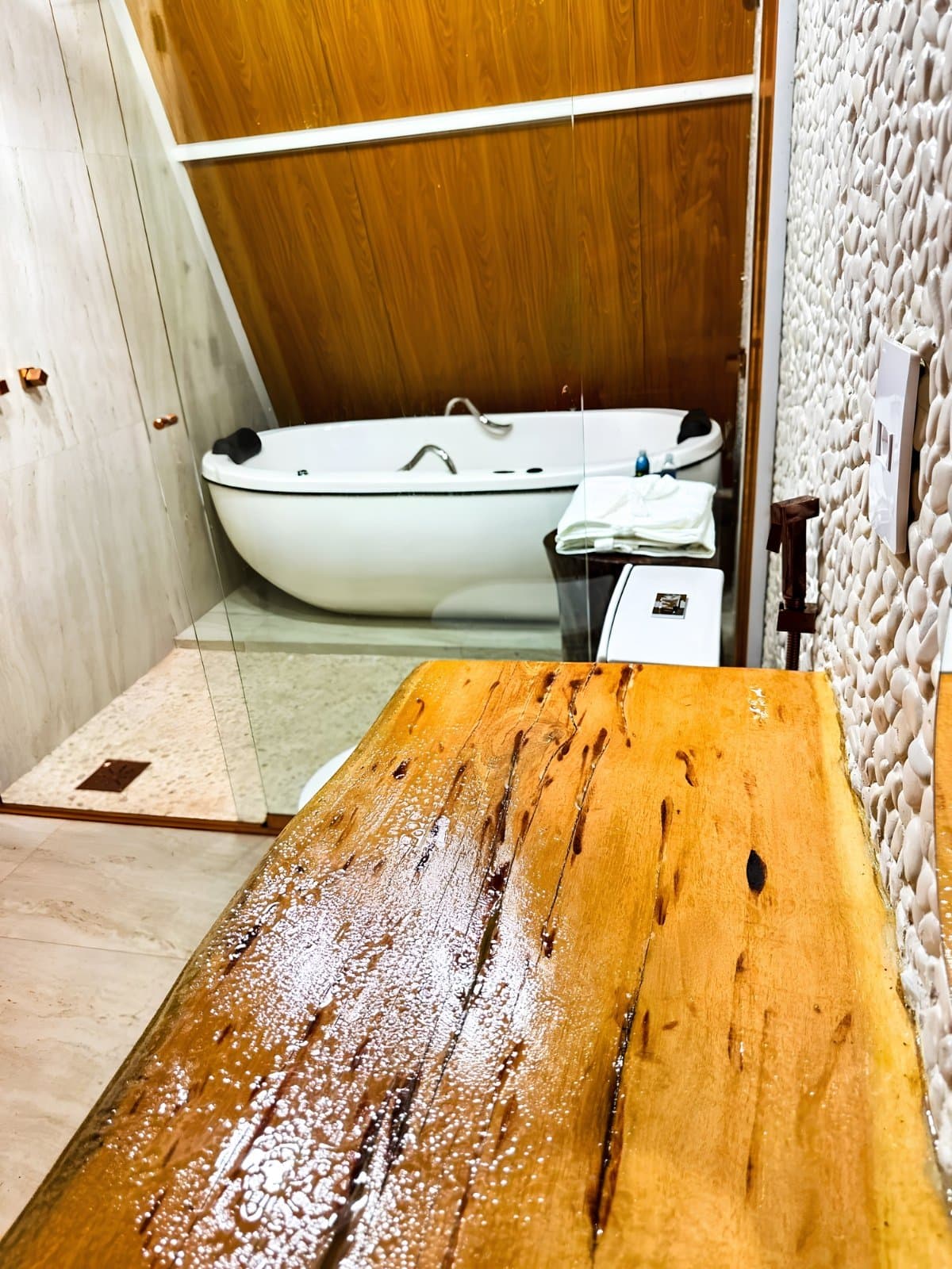 Dust off, rinse off, soak. The lodge bathroom earns its moment. A wet live-edge wooden bench in the foreground leads the eye to a freestanding oval soaking tub backed by warm wood-panelled walls and a textured white stone accent wall in a guesthouse bathroom on a jalapão trip