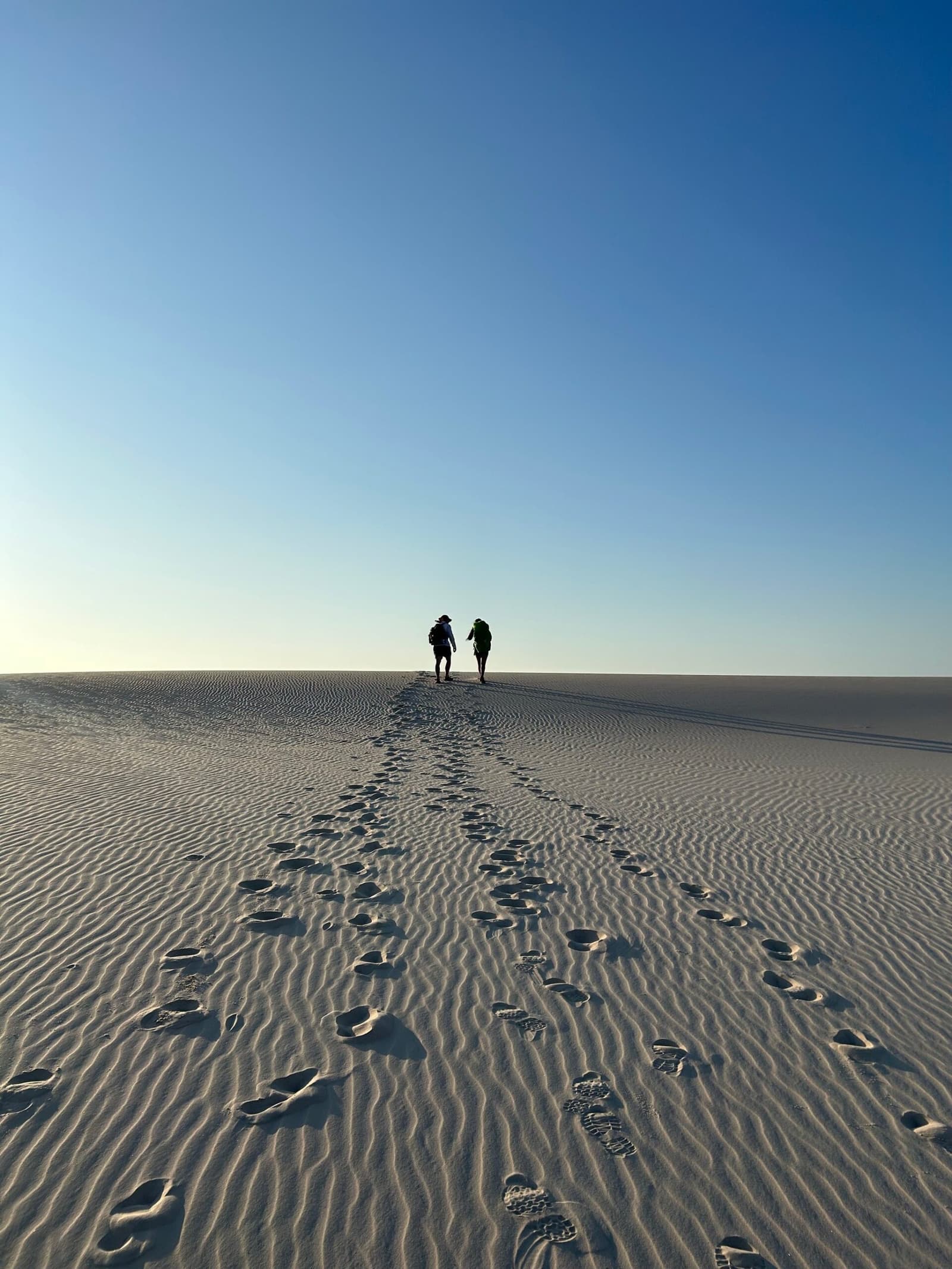 Your footprints in the sand, the horizon ahead, nothing else needed. Two hikers with backpacks silhouetted at the crest of a vast white sand dune on the Lençóis Maranhenses hike, a trail of footprints stretching back across the rippled sand beneath a clear blue sky