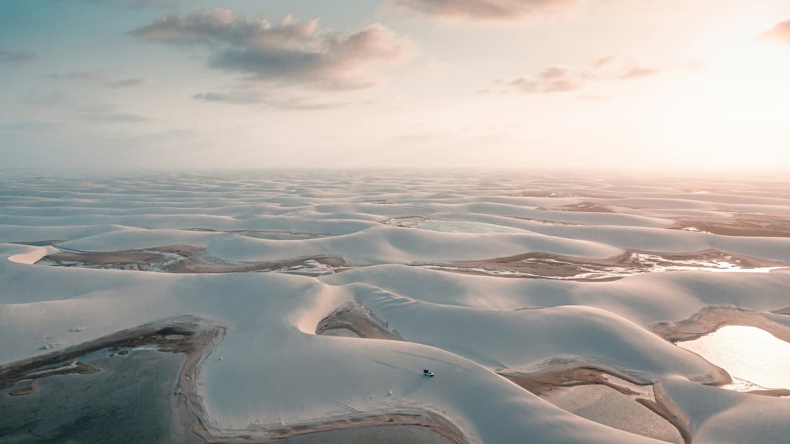 Endless white dunes, a handful of lagoons, and one very small truck putting it all in perspective. Aerial view of the Lençóis Maranhenses Trek from Atins showing a lone 4x4 vehicle crossing an endless expanse of white sand dunes dotted with shallow lagoons under a warm golden sky