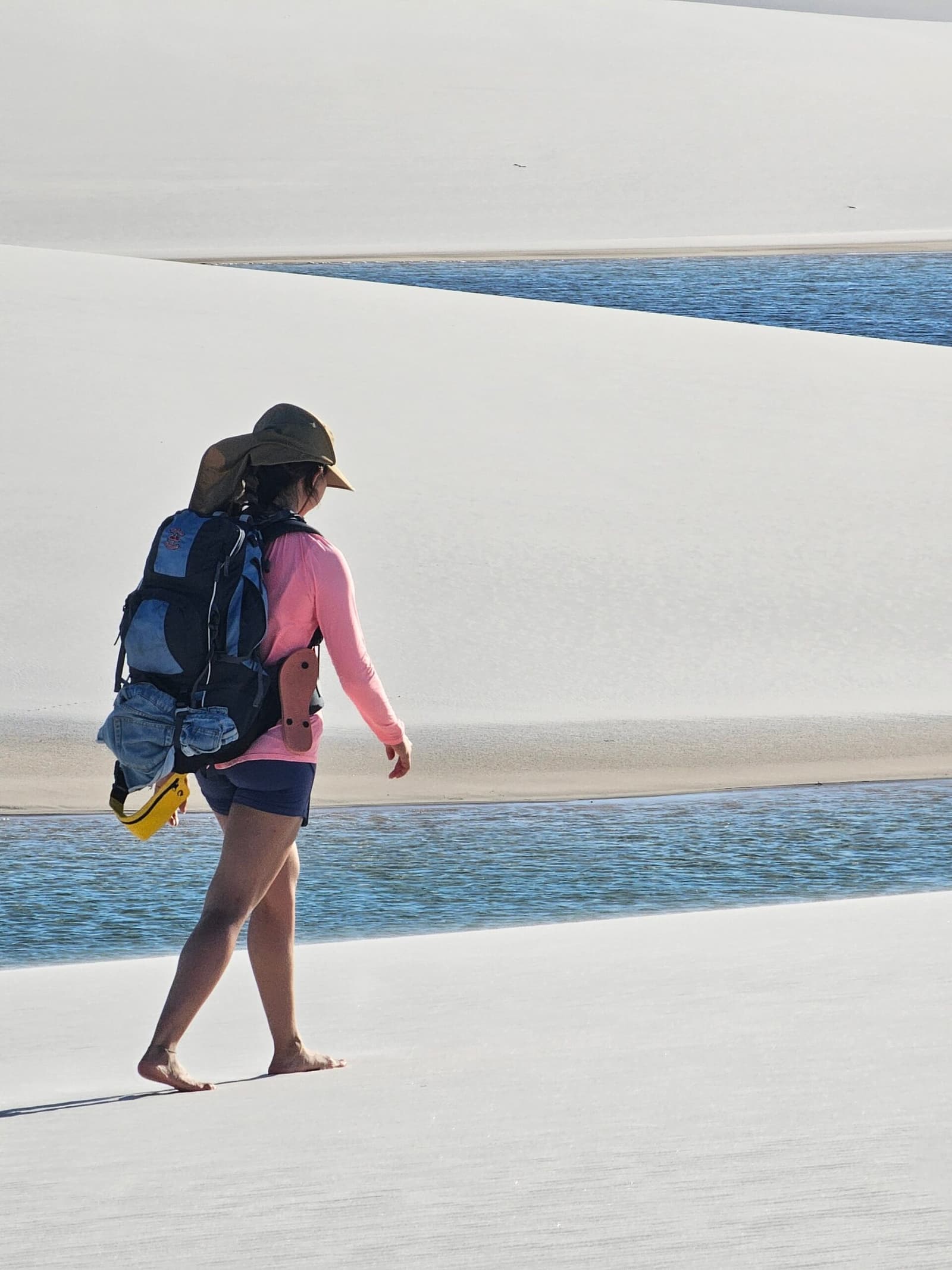 Shoes optional. The dunes have their own rules. A woman with a backpack and sun hat walks barefoot across white sand dunes beside a blue freshwater lagoon on the Lençóis Maranhenses trek from Atins