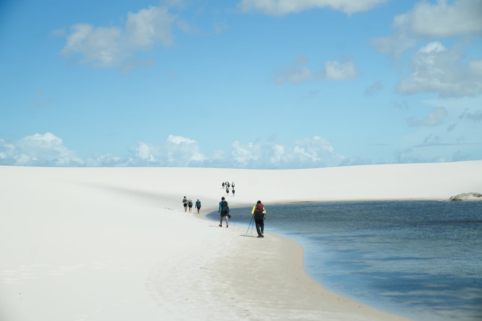 One foot in the dune, one foot in the water, that's the whole route. A line of trekkers with backpacks walks along the narrow white sand strip between a calm blue lagoon and a sweeping dune during a Lençóis Maranhenses trekking expedition under a partly cloudy sky