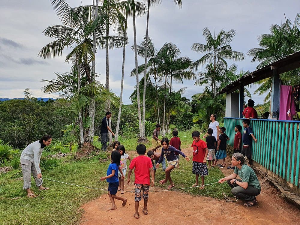 Turns out a rope and a clearing are all you need to break the ice. Children and adult visitors play a rope game together in an open dirt clearing beside a turquoise wooden building, surrounded by tall Amazonian palms, during the Pico da Neblina Expedition community visit.
