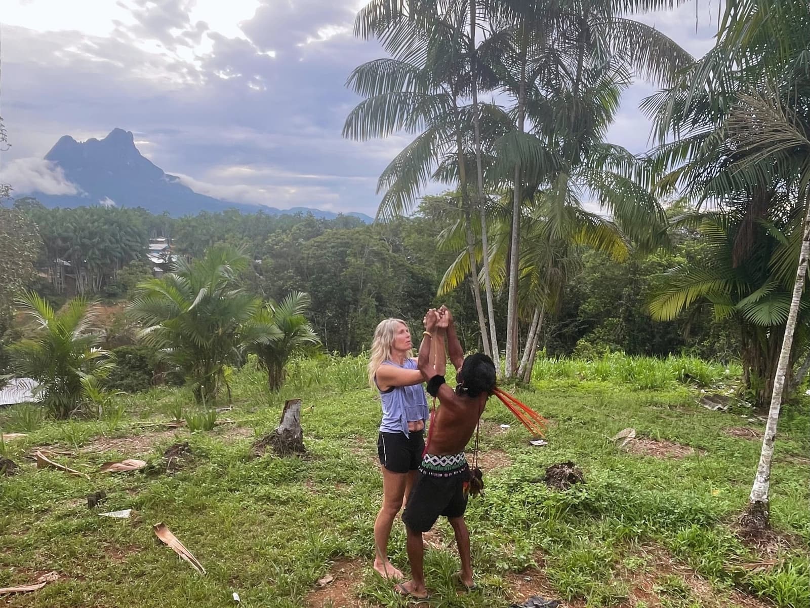 Hands raised, the forest witnesses. The trek begins the right way. A traveler and a Yanomami elder raise their joined hands during a traditional blessing ceremony before the Pico da Neblina Expedition, with palm trees and a dramatic peak rising through clouds in the background.