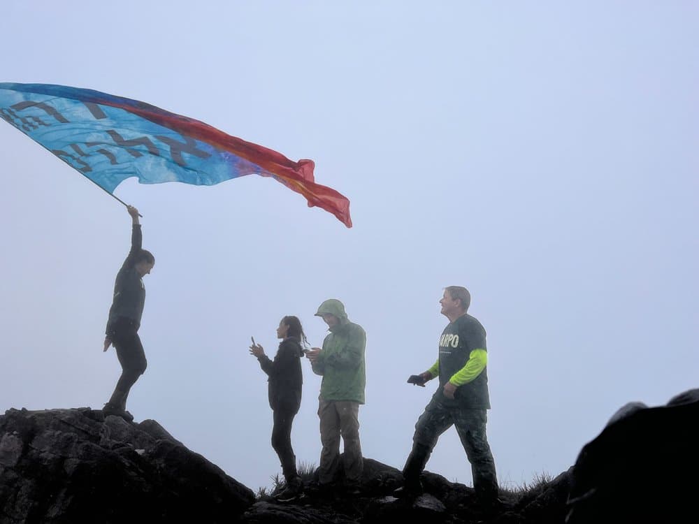 The flag goes up, the clouds close in, and nobody minds one bit. Four hikers silhouetted against thick mist on a rocky ridge during a Pico da Neblina Expedition, one person raising a large colorful flag that billows in the wind