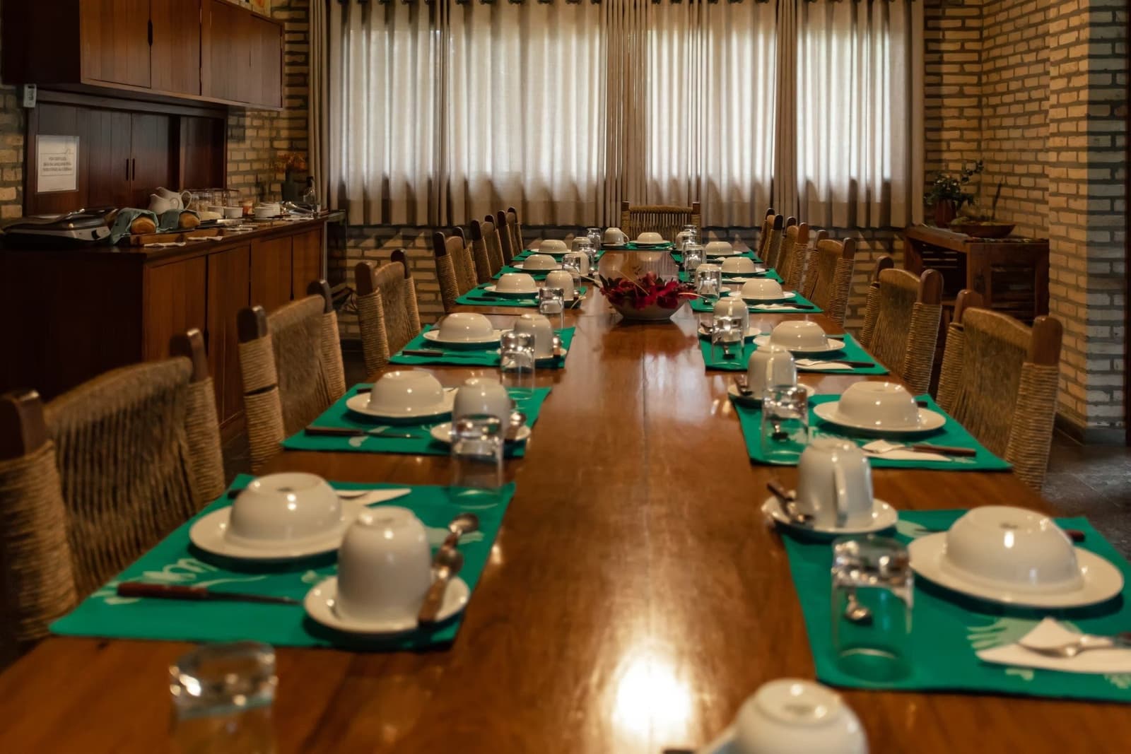 Full board, proper table settings, and a long day of wetlands ahead. Long wooden dining table set for a group with white dome-covered bowls, green placemats, and woven chairs inside a brick-walled lodge dining room on a southern pantanal safari