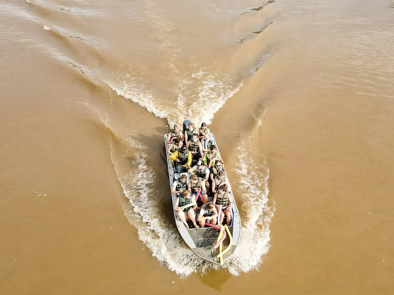 Full boat, open water, and the lodge still hours away. Good. Aerial view of a packed speedboat cutting through muddy brown river water on a southern pantanal safari, passengers wearing life jackets with a wide V-shaped wake behind them