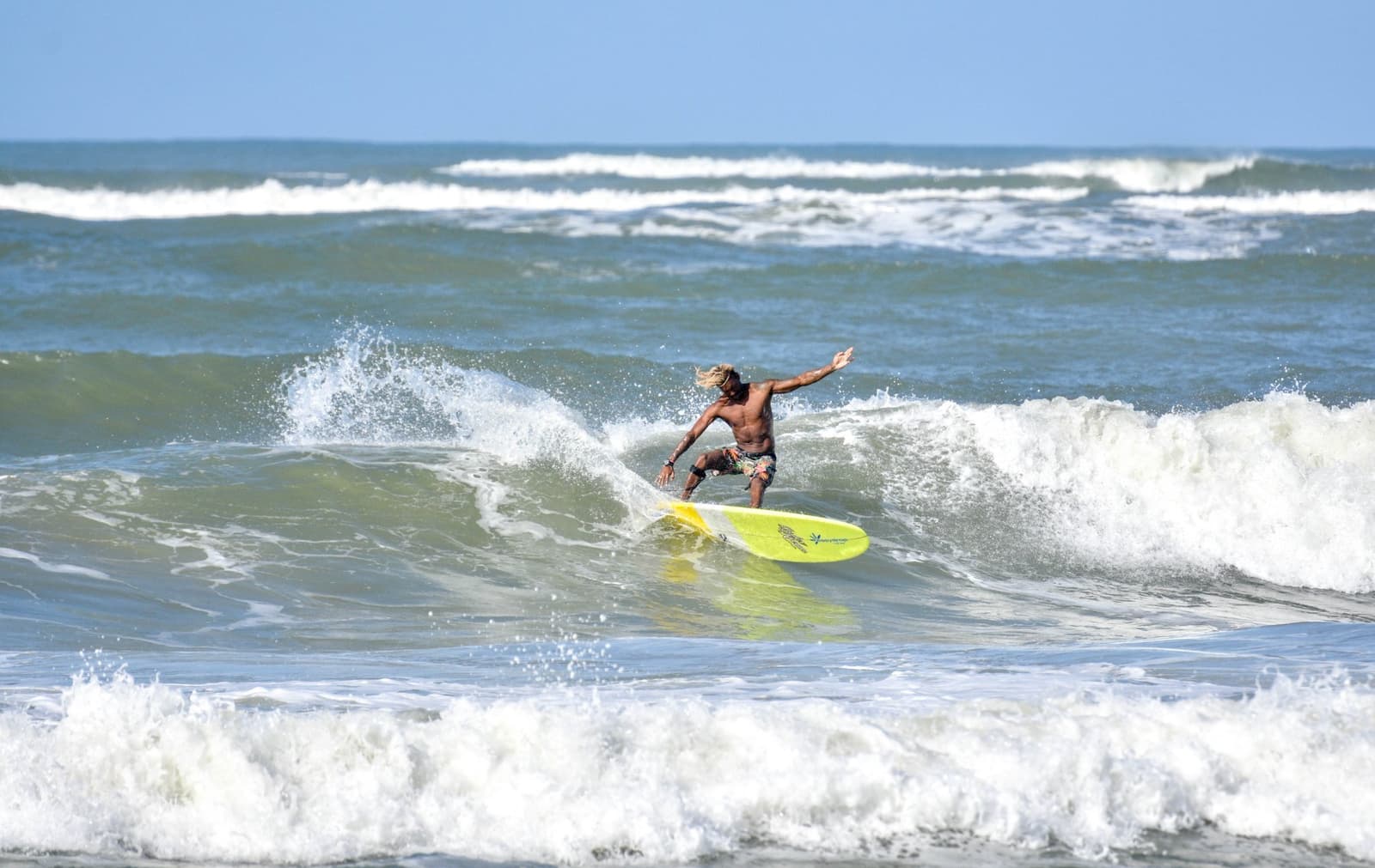 Arms wide, hair flying, completely in it. That's the goal. Photo: Picasa A surfer with long blond hair rides a yellow longboard through a breaking wave during surf lessons in Itacaré, arms outstretched for balance under a clear blue sky.
