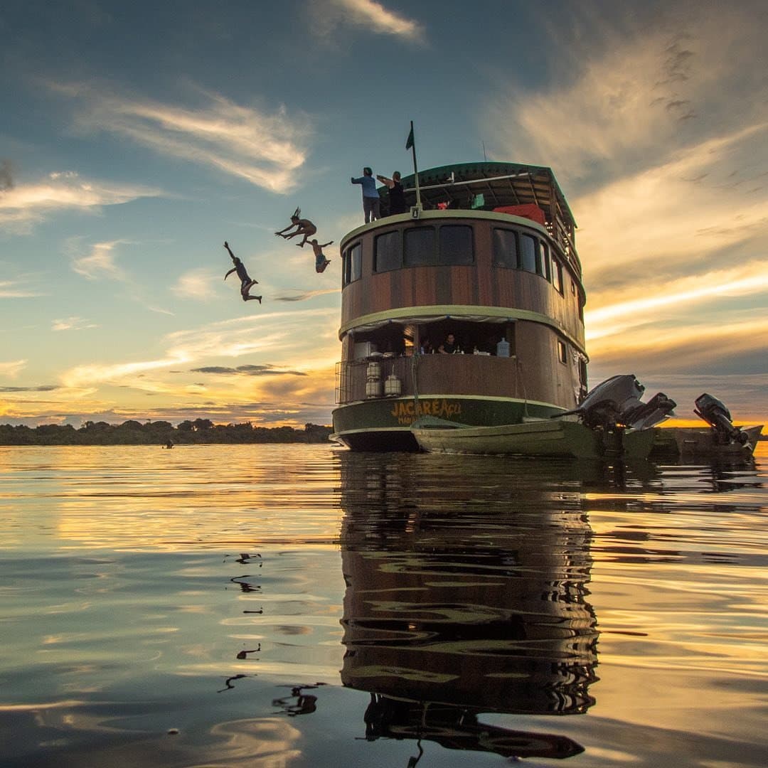 Some travel memories happen mid-air, somewhere between the top deck and the river. Guests leaping from the upper deck of the Jacaré-Açu into a glassy river at golden hour on an Amazon cruise trip in Brazil, their silhouettes frozen mid-air against a vivid sunset sky