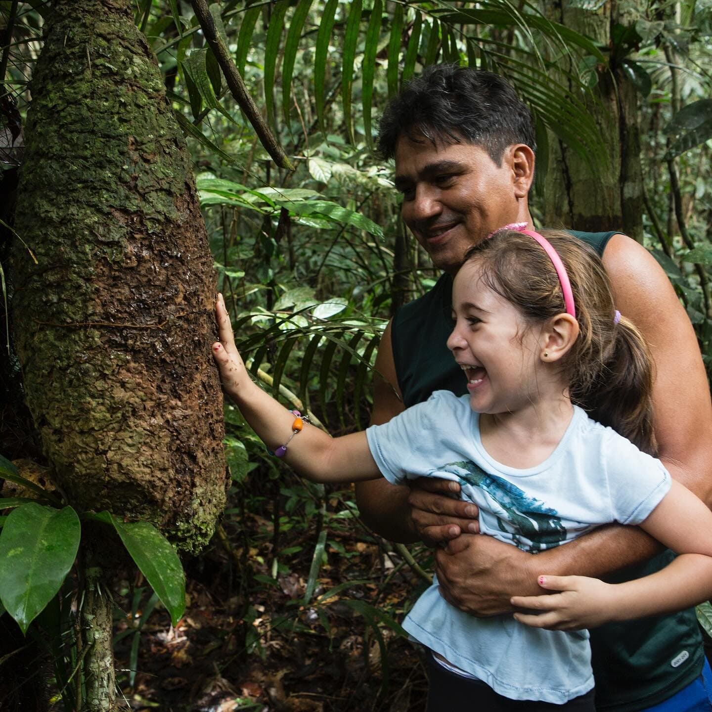 That laugh is the forest saying hello back. A smiling man holds a laughing young girl as she touches the textured bark of a large rainforest tree during an amazon river cruise brazil forest hike, dense green vegetation surrounding them