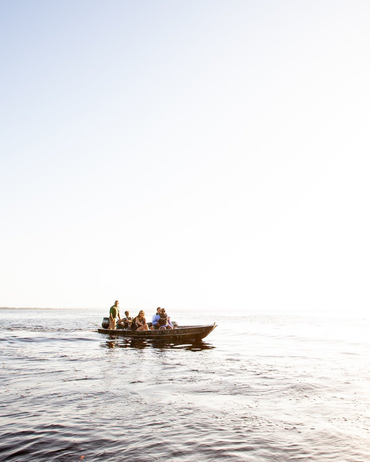 Five people, one boat, and a whole lot of river to themselves. A small wooden motorboat carrying five passengers drifts across the calm, dark waters of an Amazon river cruise in Brazil, under a bright hazy sky with no land in sight.