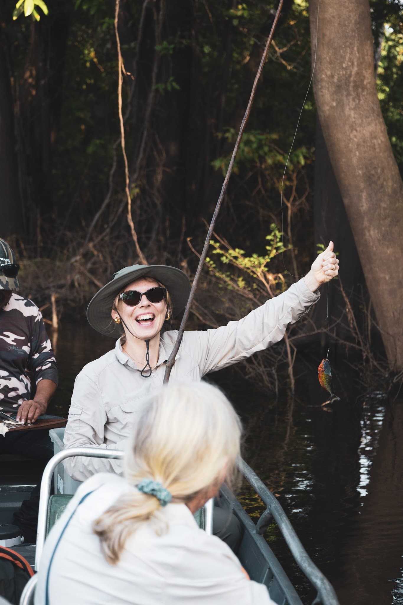 First catch, biggest grin. Piranha fishing earns it. A laughing woman in a sun hat holds up a small caught fish from a motorized canoe during an amazon turtle lodge piranha fishing excursion on a dark jungle waterway