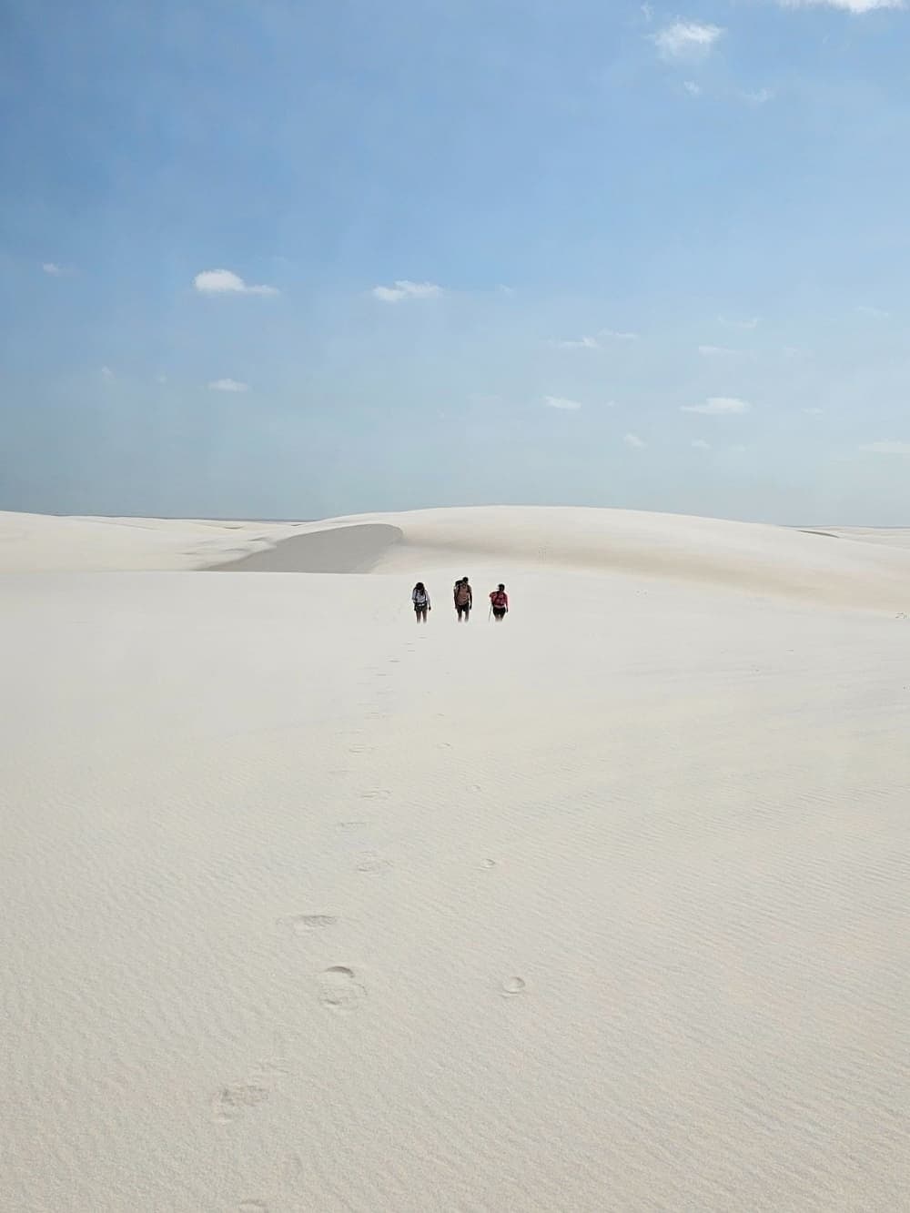Three people, an ocean of white sand, and a long way to go. Three hikers with backpacks walking across vast white sand dunes on the Lençóis Maranhenses hike, their footprints trailing behind them under a pale blue sky