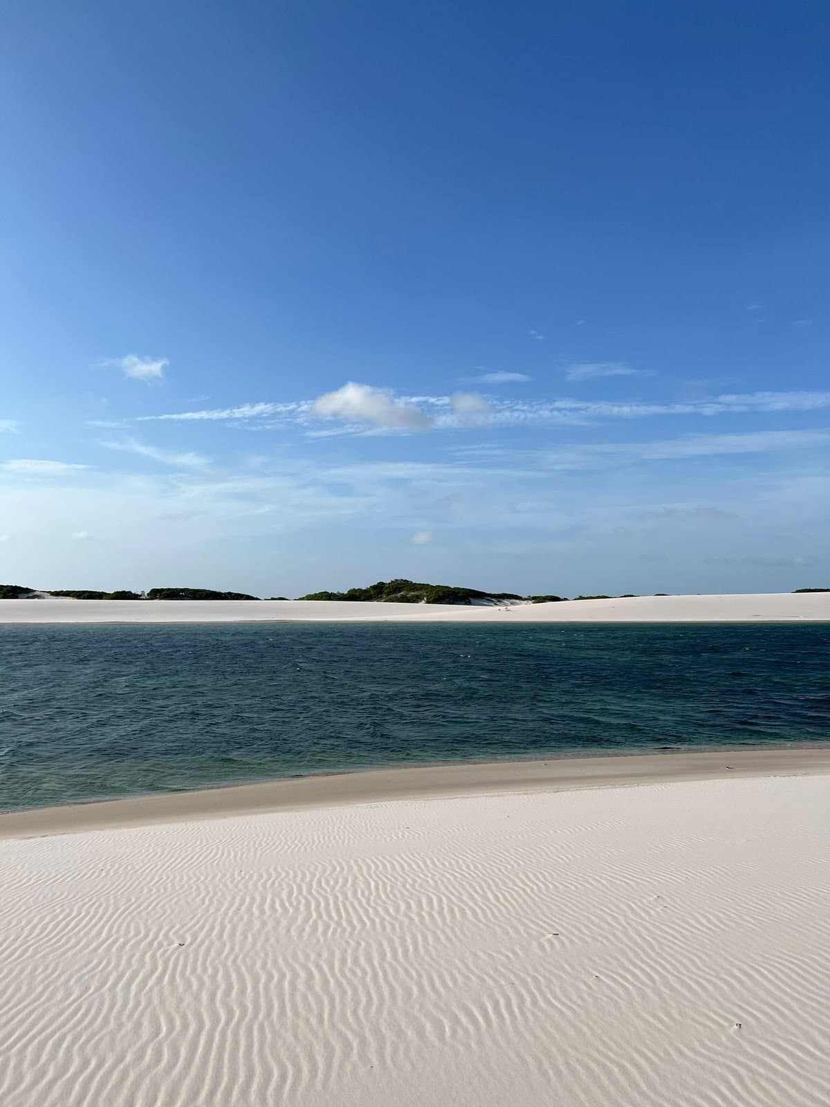 Sand this white should come with a warning label. Wide freshwater lagoon flanked by rippled white sand dunes under a clear blue sky in Lençóis Maranhenses Santo Amaro, with scattered green vegetation on the far dune ridge