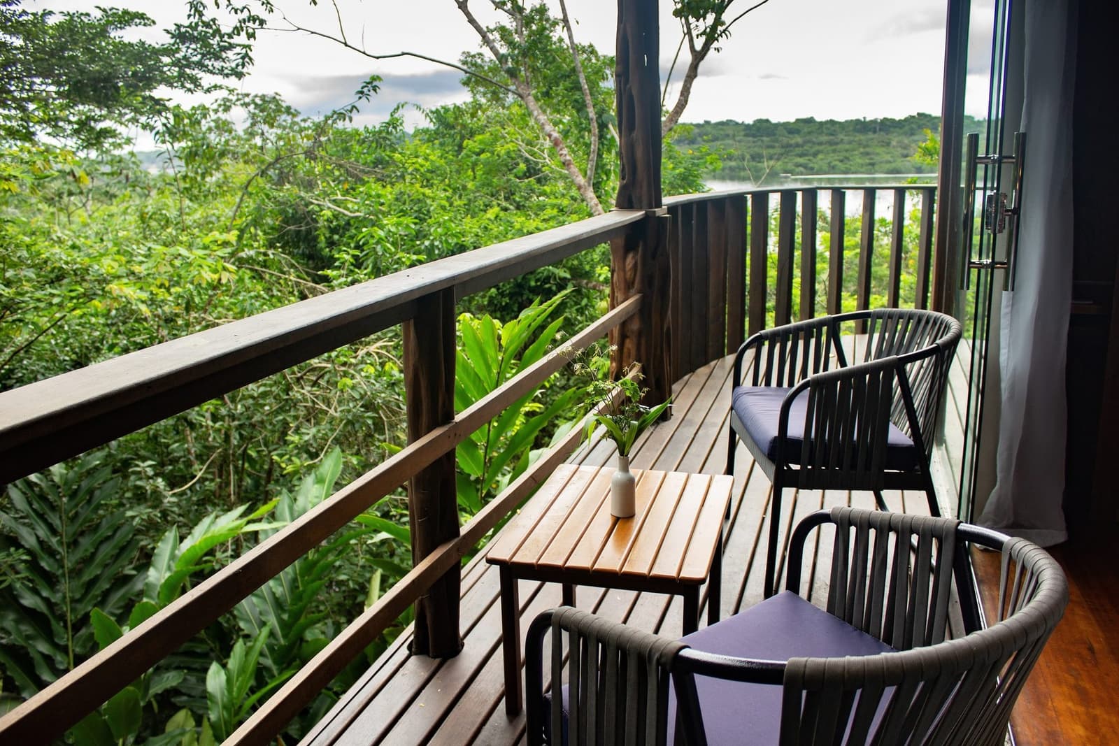 Your morning coffee just got a serious upgrade. A curved wooden balcony at Mirante do Gavião fitted with two cushioned chairs and a small slatted table overlooking a dense green rainforest canopy and a river glimpsed in the distance