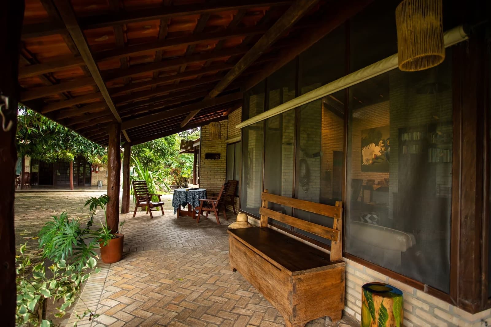 Sit down, take your boots off. The veranda has been waiting. Covered brick veranda of a southern pantanal safari lodge, with a rustic wooden bench and chest in the foreground, wooden chairs around a table mid-corridor, and a lush garden courtyard beyond