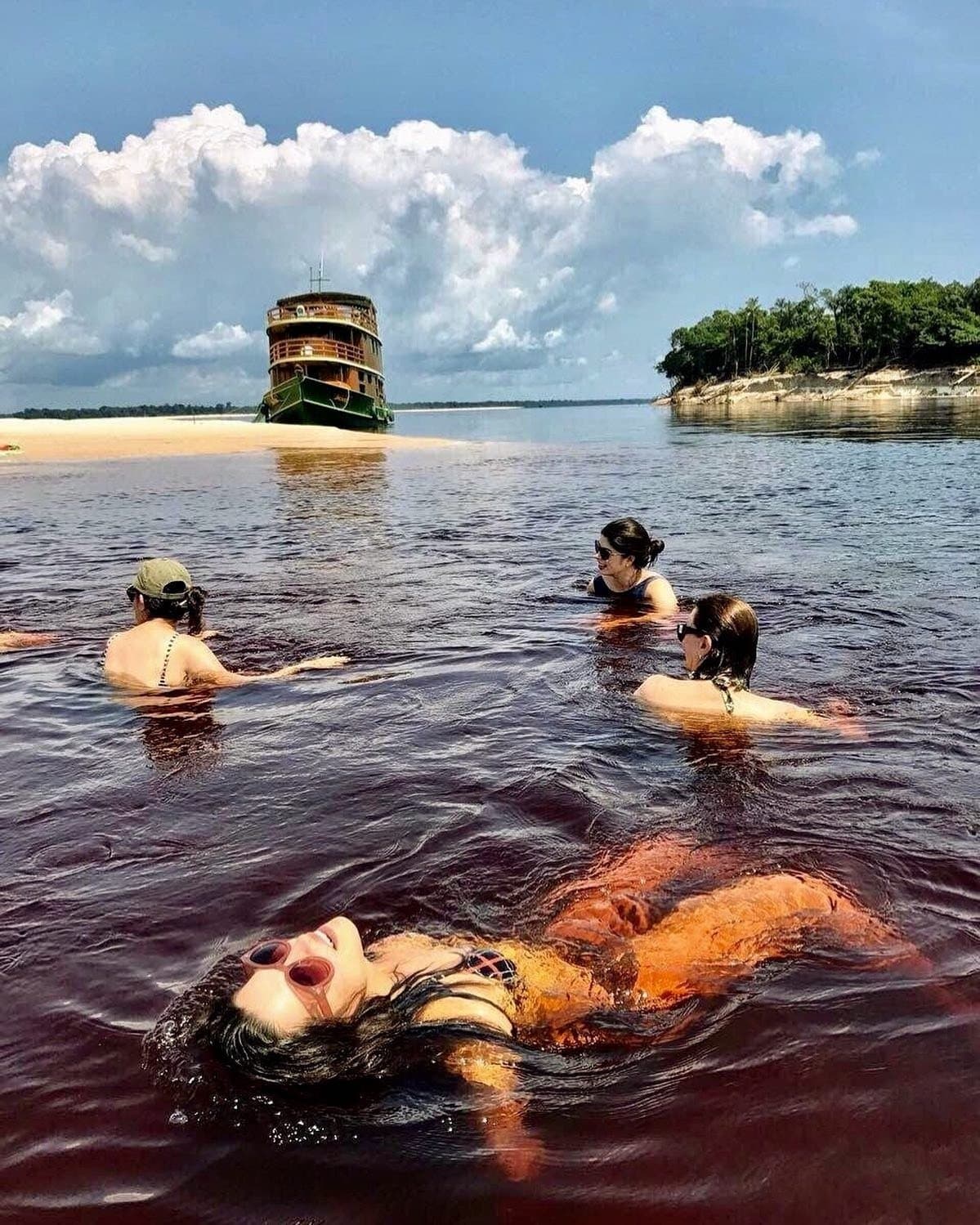The water is warm, dark as tea, and yes, you do get in. Four travelers swimming in the dark tannin-stained waters of the Rio Negro on an Amazon cruise trip in Brazil, with a classic multi-deck wooden riverboat anchored near a white sandbar and forested riverbank under a cloudy sky