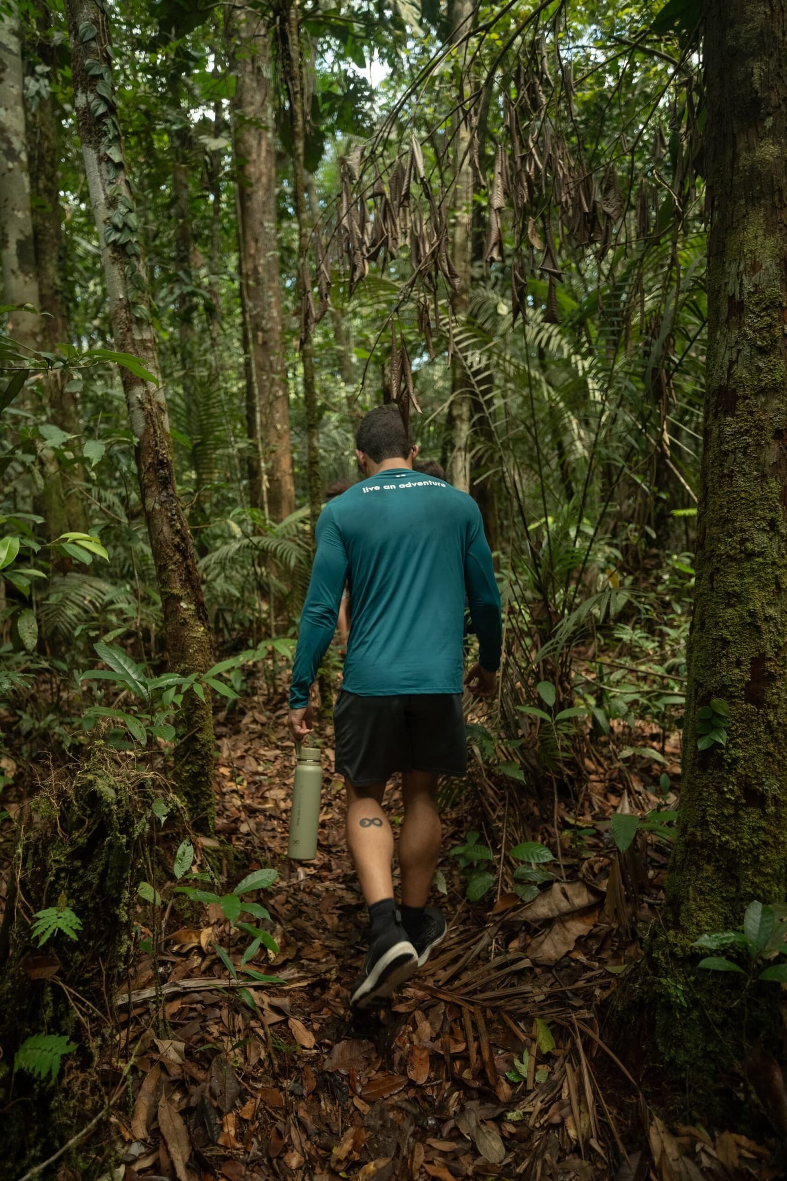Good boots, a full bottle, and a trail that keeps its secrets close. A hiker in a teal long-sleeve shirt carrying a water bottle walks away from the camera along a narrow leaf-covered trail through dense rainforest on an amazon river cruise brazil excursion.