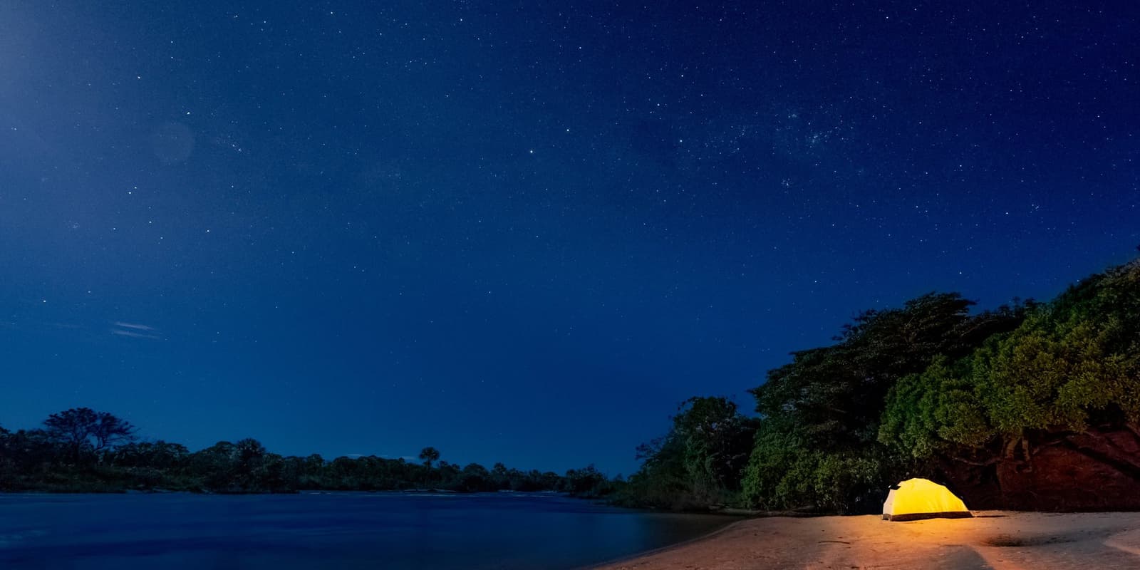 One small light, one enormous sky, and the river keeping you company all night. Photo: Alberto Andrich A glowing yellow tent pitched on a sandy river beach during a Jalapão trekking expedition, under a deep blue starlit sky with dense cerrado forest lining the opposite bank