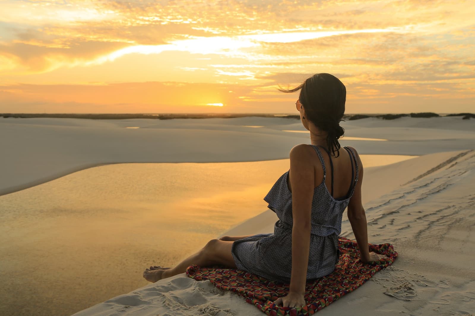 Some views earn a quiet sit-down. This is one of them. A woman sits on a colorful blanket atop a white sand dune in Lençóis Maranhenses Full Itinerary, gazing at a vivid golden sunset over the vast dune landscape