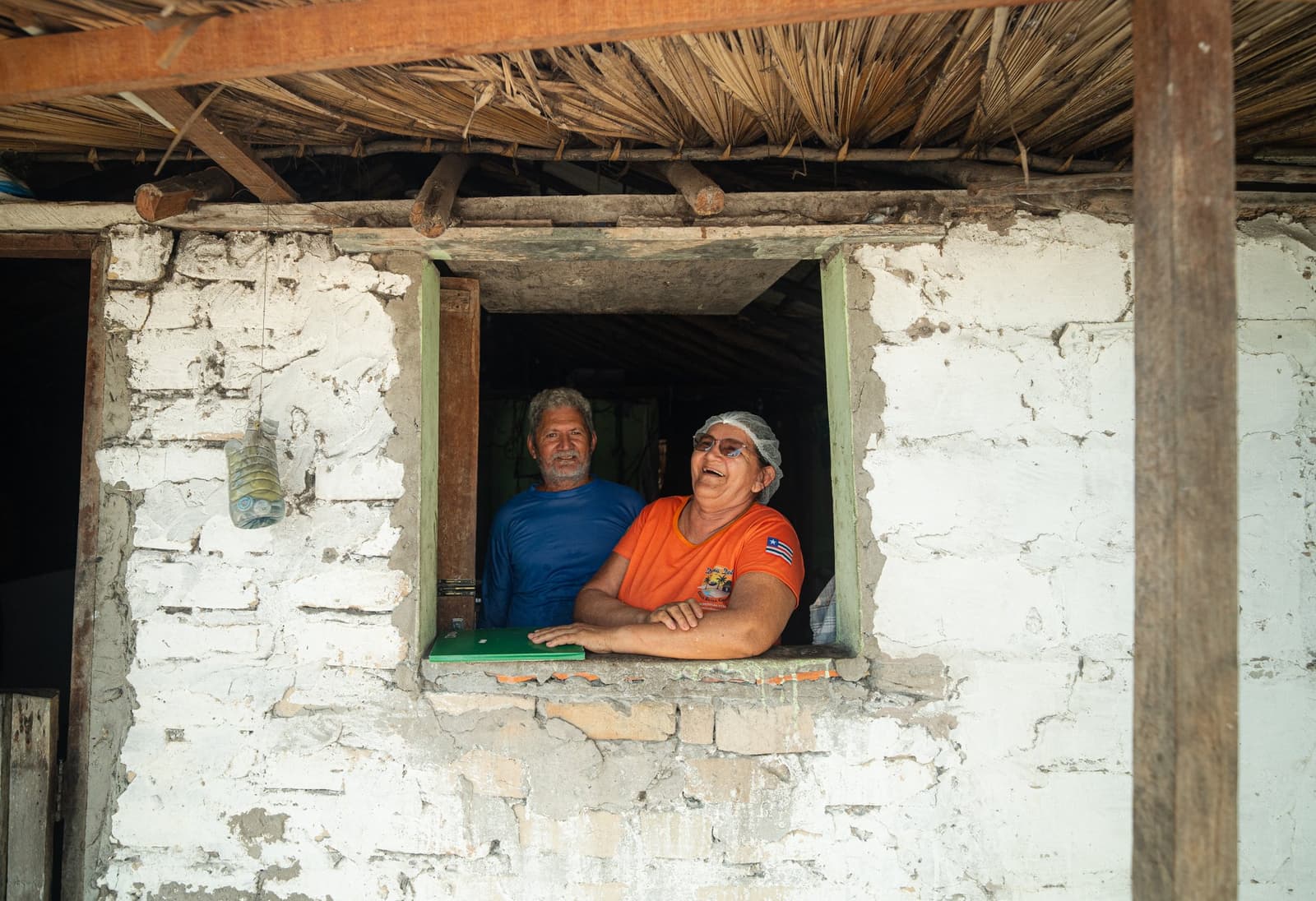 The people who live here are half the reason to come. A smiling woman in an orange shirt and hairnet and a man in a blue shirt lean out of a whitewashed stone window beneath a thatched roof, welcoming visitors on the Lençóis Maranhenses hike.