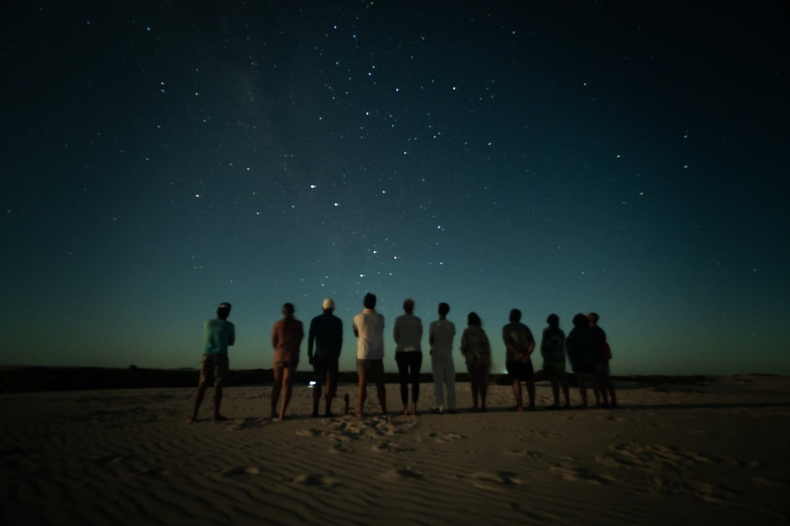 No screens, no noise, just ten people and a sky full of stars. A group of travellers stands in a row on white sand dunes during a Lençóis Maranhenses trek from Atins, silhouetted against a deep blue night sky filled with stars and a faint Milky Way