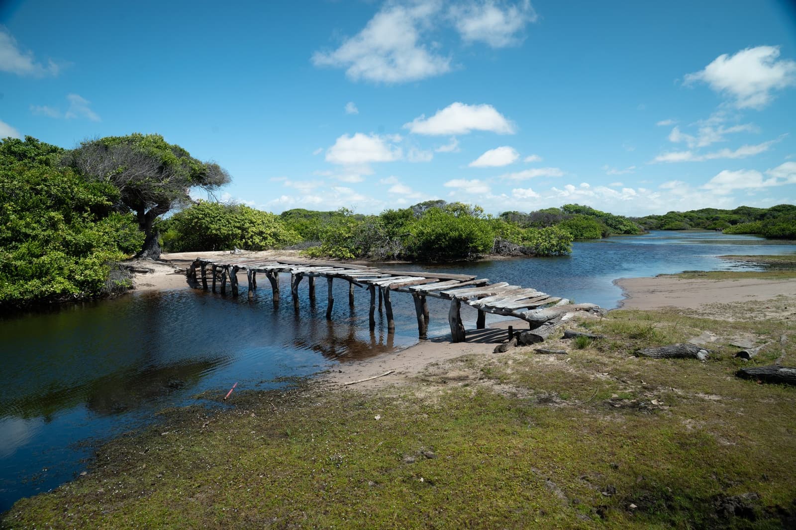 Built to last, repaired with optimism, loved by the landscape either way. A weathered wooden footbridge crosses a shallow tidal channel bordered by low coastal shrubs and sandy banks on the lencois maranhenses trek route under a blue sky with scattered clouds.
