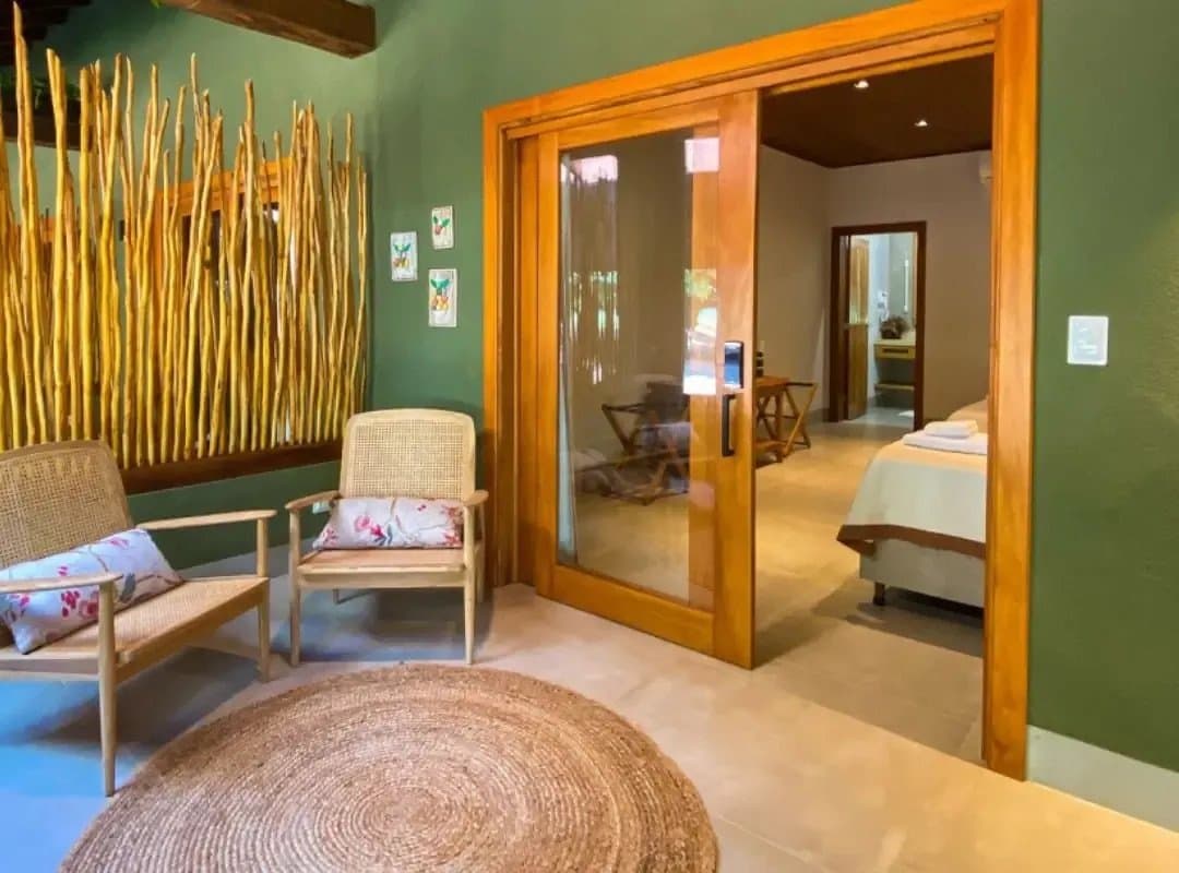 Rustic materials, a green palette, and enough quiet to hear yourself think. Sitting area of a southern pantanal safari lodge featuring rattan chairs, a round woven rug, botanical art prints, a decorative wall of golden dried branches, and a sliding glass door opening into a bedroom with a neatly made bed