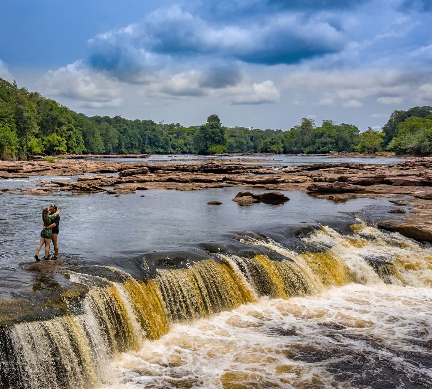 Some moments you don't plan, the river just arranges them for you. A couple embracing on a rocky ledge beside a wide cascading waterfall on an Amazon jungle cruise Brazil itinerary, surrounded by dense tropical forest under a dramatic cloudy sky