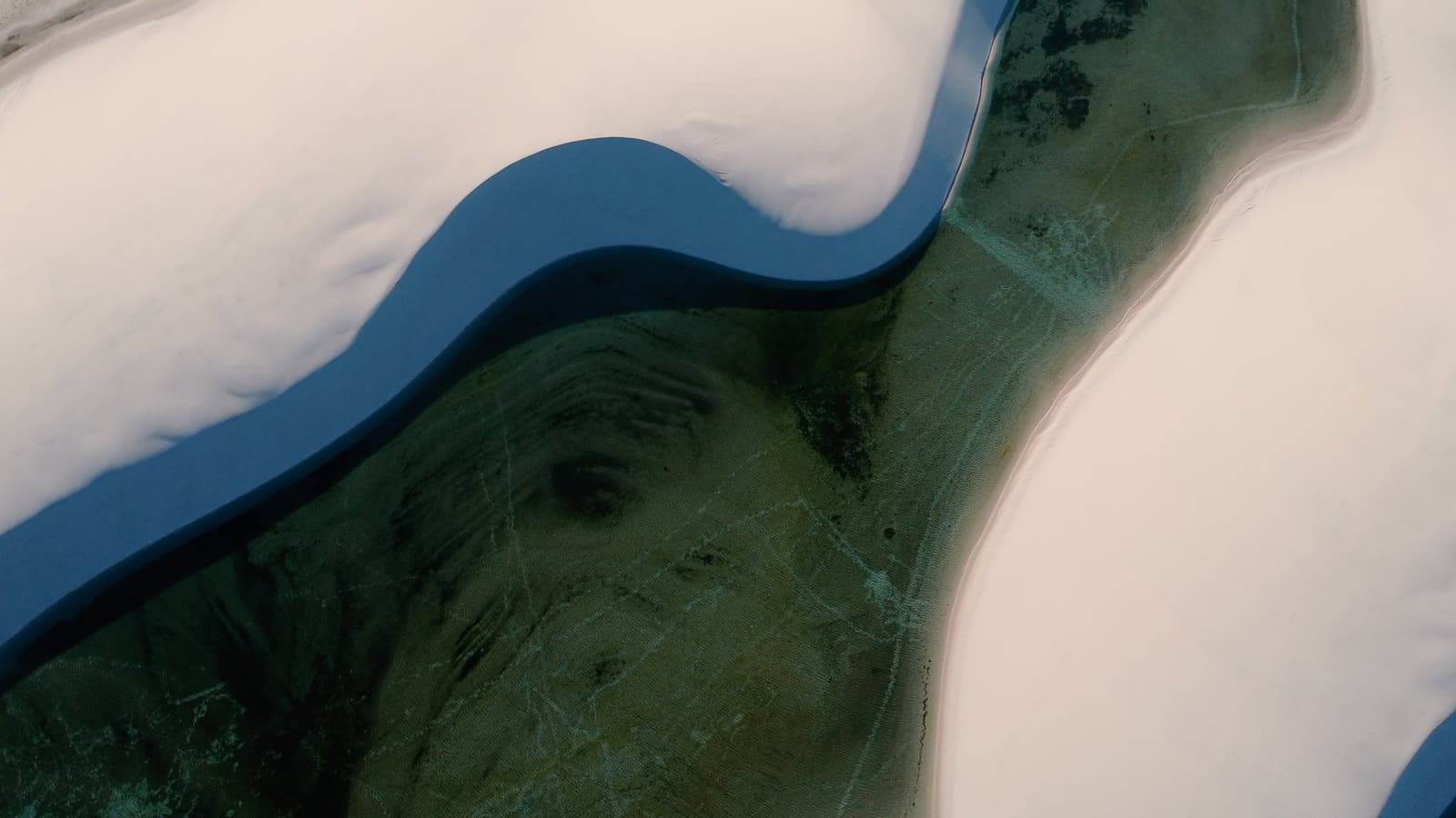 From above, the dunes look like they are slowly swallowing the land whole. Aerial view of a deep blue lagoon cutting through white sand dunes during the Lençóis Maranhenses hike, with green aquatic vegetation visible through the clear water