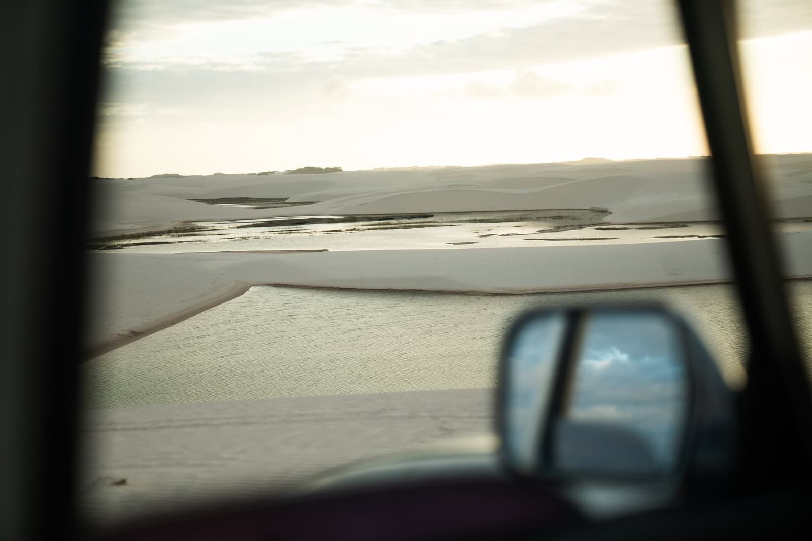 The dunes arrive before you do, seen first through glass, already vast. View through a 4x4 vehicle window of white sand dunes and shallow green lagoons stretching to the horizon during the Lençóis Maranhenses trek from Atins, with the side mirror reflecting an overcast sky