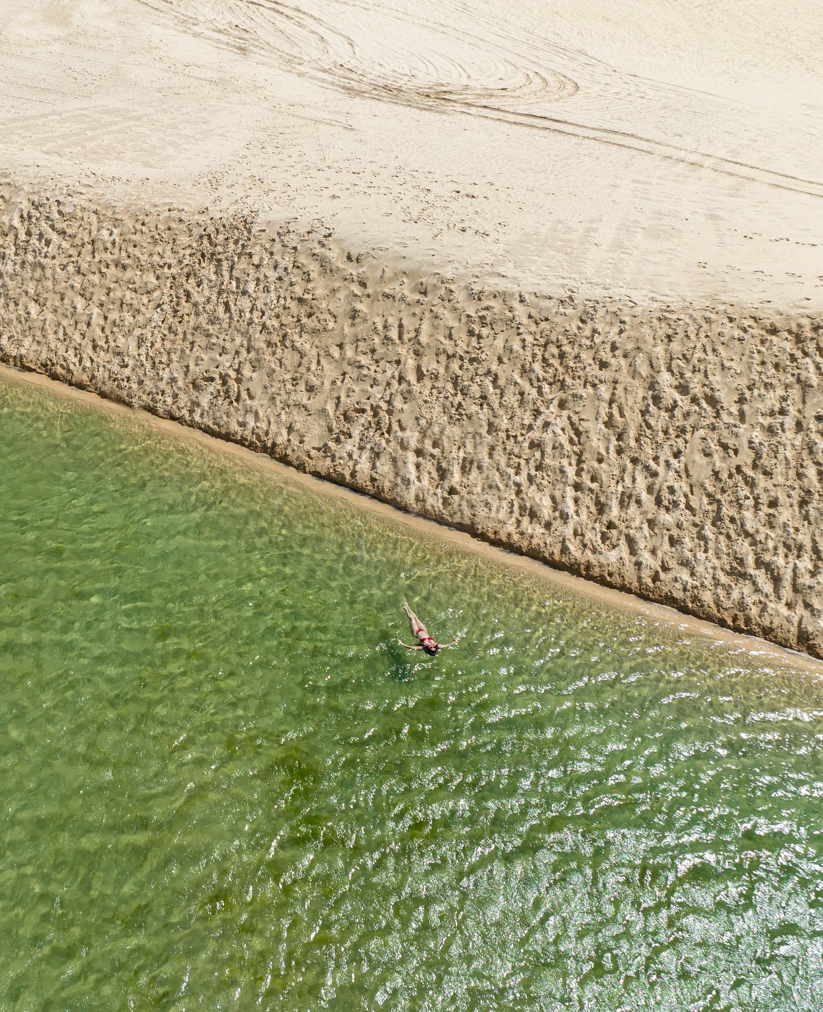 Just you, the green water, and a sandbar that has no plans for the afternoon. Aerial view of a lone swimmer floating in clear green water beside a textured sandbar on the Lençóis Maranhenses trek from Atins