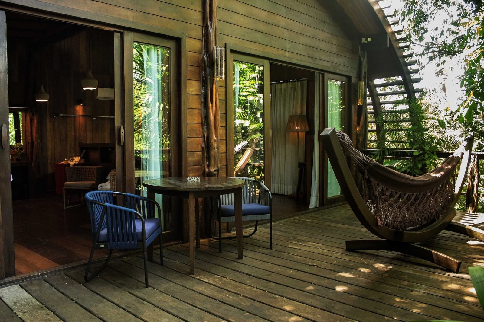 Your afternoon nap spot has already been decided for you. A timber deck at Mirante do Gavião features a round table with two cushioned chairs, a woven hammock on a curved stand, and sliding glass doors opening into a warmly lit lodge room surrounded by dense rainforest foliage