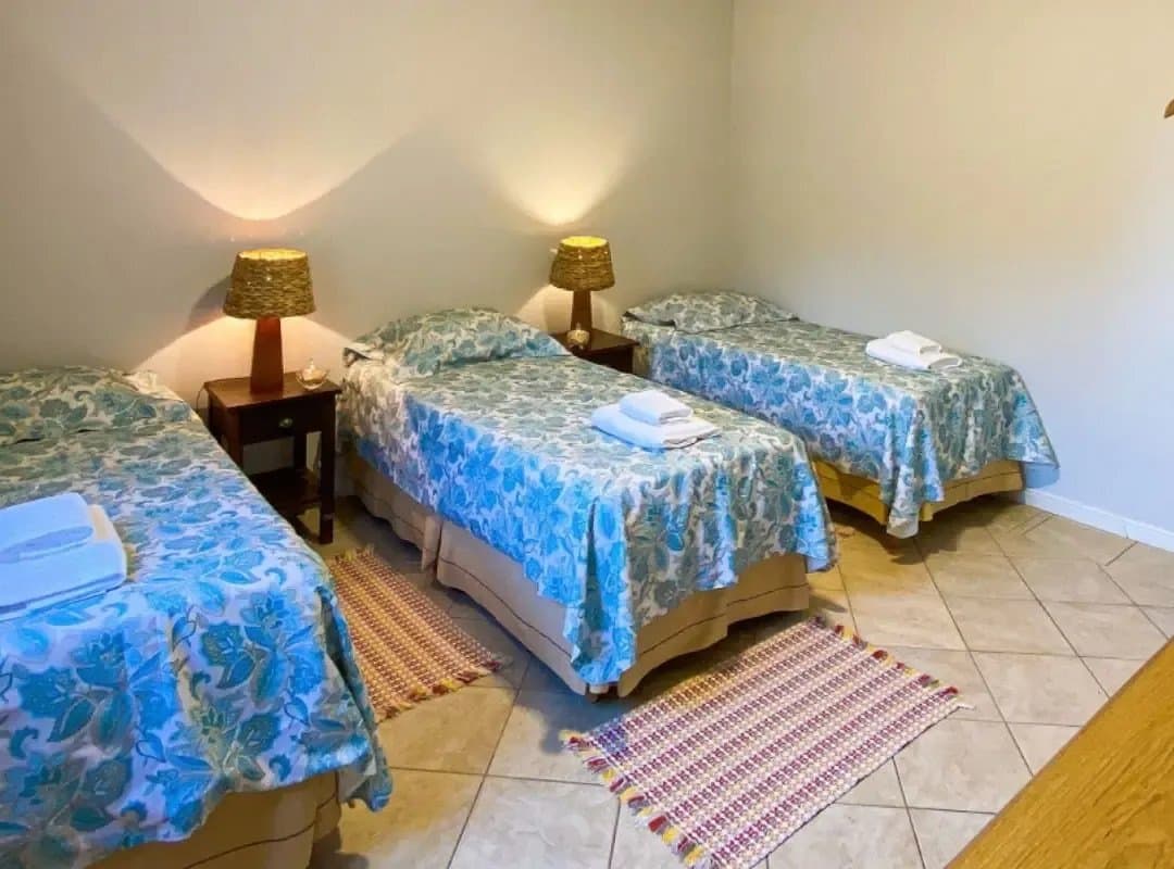 Simple, tidy, and ready for you after a long day on the floodplain. Three single beds with blue floral bedspreads, folded towels, woven lamp shades, and colourful rugs on a tiled floor in a southern pantanal safari lodge room
